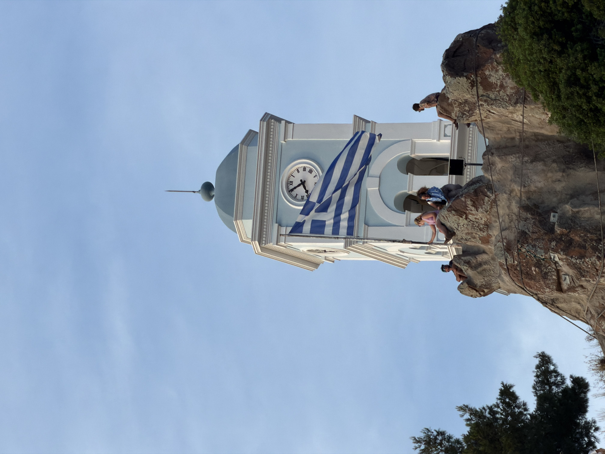 This image, set in Poros, Greece, features a striking light blue and white clock tower against a clear, pale blue sky. The tower has a distinct domed top with a finial. Below the dome, a white clock face with Roman numerals is visible, showing the time to be approximately 4:50. A large Greek flag, adorned with its characteristic blue and white stripes and a white cross in the canton, billows in front of the clock face, indicating a gentle breeze.
To the right of the clock tower, a rugged, multi-toned rocky cliff or outcrop dominates the foreground, with patches of green foliage and trees growing on its surfaces. Several individuals are present on this rocky terrain. One person is seen higher up on the cliff face, while another with dark hair is seated further down. A third person, wearing a pink top, is positioned closer to the base of the rock, near the tower. The people appear to be casually observing their surroundings or resting.
The scene conveys a bright, sunny day, likely in the late afternoon, with the individuals enjoying the outdoor environment and the panoramic views from their elevated position. The architectural style of the tower, the national flag, and the natural rocky landscape strongly suggest a Mediterranean, specifically Greek, island setting.