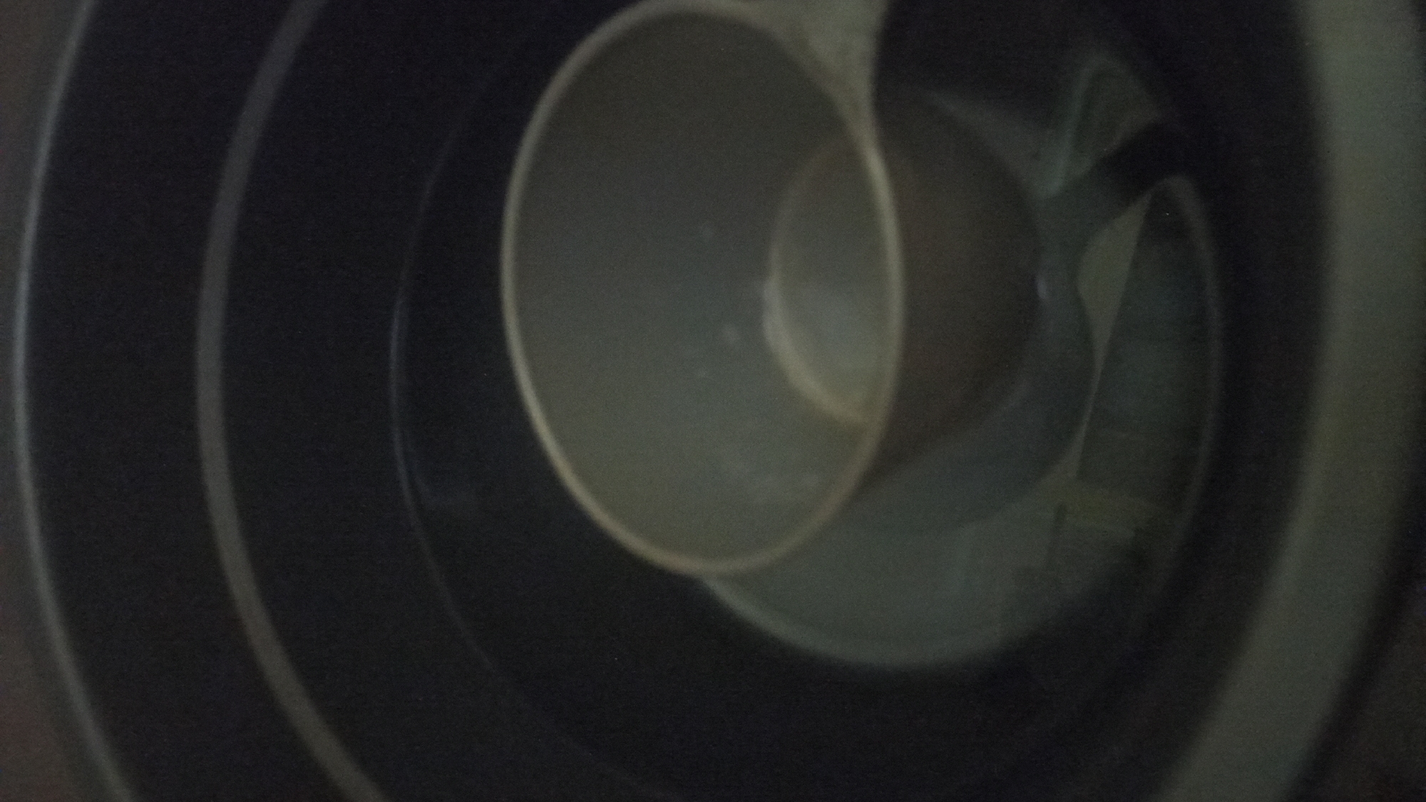 A close-up, dimly lit image shows a grey plastic measuring cup partially submerged in water within a larger, dark grey plastic container. The container has concentric ridges visible on its interior surface. The measuring cup is upright, with its handle facing towards the right side of the frame. Some faint reflections are visible on the surface of the water. The image appears to be taken indoors, in a non-public setting, with no discernable location in Tuba, Indonesia.