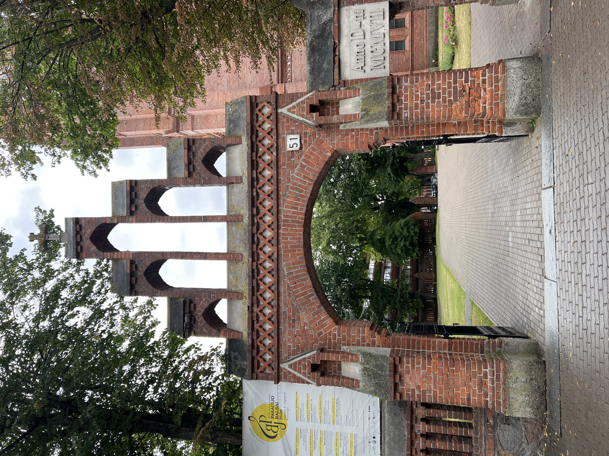 The image shows the entrance gate to Palanga Church in Palanga, Lithuania. The gate is made of red brick and features a Gothic architectural style with arched openings and decorative brickwork. Above the main arch, there is a series of smaller arched openings culminating in a cross at the very top. To the left of the gate, a large banner with text and graphics is displayed. To the right of the gate, a stone plaque is embedded in the brick wall with the inscription "Anno D-m MCMVIII". Beyond the gate, a paved walkway leads into a green lawn area with trees and the church building visible in the background. The weather appears to be overcast.