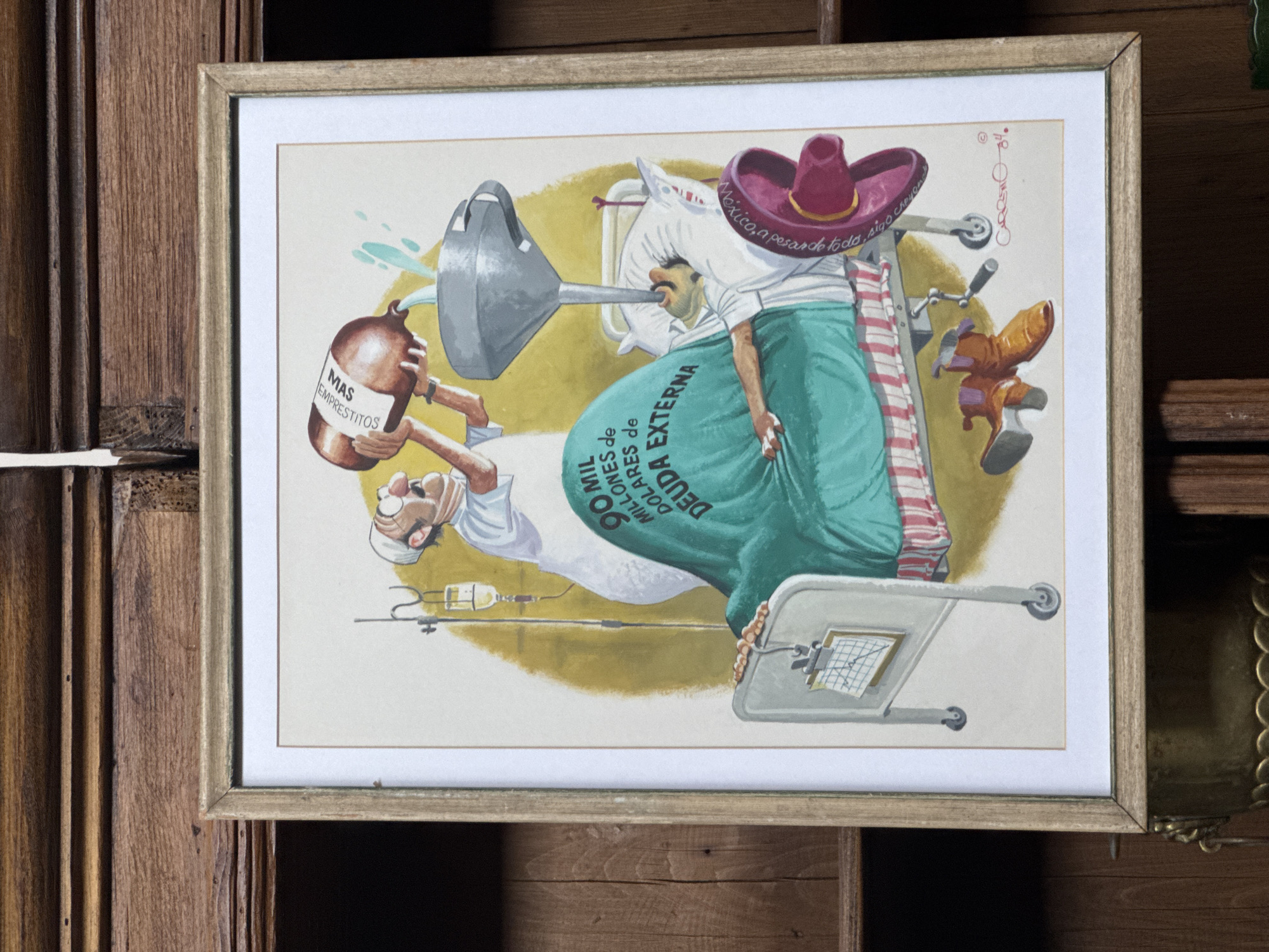 The image shows a framed political cartoon depicting Mexico's debt. A person wearing a sombrero and a green bag labeled "90 Mil Millones de Dolares de Deuda Externa" (90 Billion Dollars of External Debt) is being filled with liquid from a bottle labeled "Mas Emprestito" (More Loans) via a funnel, lying on a hospital bed. The scene takes place within a wooden shelf, adding context to the cartoon's setting. There is no information available about the city of San Miguel de Allende, Mexico, in the image.