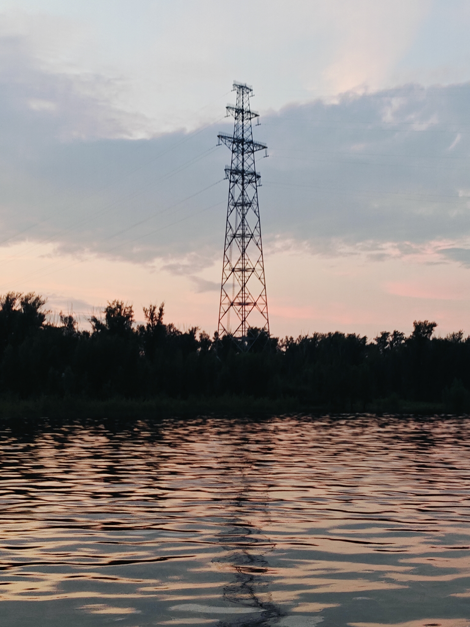 The image depicts a tall, lattice-like metal electricity pylon standing prominently against a dusk sky over a dark line of trees. The sky exhibits a gradient of soft colors, transitioning from a pale blue at the top to hints of pink and peach near the horizon. The water in the foreground reflects these sky colors and the dark silhouette of the trees, creating a textured, rippling surface. The scene appears to be a natural landscape with man-made infrastructure. No people or animals are visible in the image. The location is identified as Noviy, Russia.