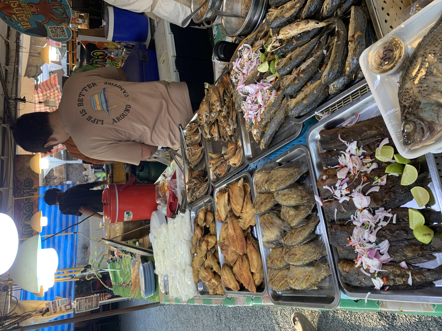 This vibrant photograph captures an outdoor market stall, likely in บ้านบางรักษ์, Thailand, specializing in various fish dishes. The scene is bustling with food displayed for sale.
In the foreground, a man, seen from behind, wears a beige t-shirt with the text "LIFE IS BETTER AROUND CAMPFIRE" encircling a graphic of a hat. He stands behind the stall, which is laden with an array of prepared fish. To his right, multiple metal trays hold different types of cooked or fried fish. Some trays feature whole fish garnished with sliced red onions, small chili pieces, and fresh lime wedges, indicating a readiness for consumption. One large whole fish, possibly a tilapia, is individually packaged on a white foam tray, wrapped in clear plastic, with a small container of dipping sauce beside it. Other trays display smaller, dried or fried fish, some appearing to be crispy.
Further back, another person is partially visible, also attending the stall near a large red cooler or water dispenser. Overhead, several round white lights illuminate the stall, suggesting it might be evening or an indoor market setting. A blue and white striped tarp or awning provides shade or cover. To the left, a sign with an octopus graphic displays Thai text, which translates to "grilled squid" (ปลาหมึกย่าง), hinting at other offerings. The overall impression is one of a lively street food or market environment, offering fresh and prepared seafood.