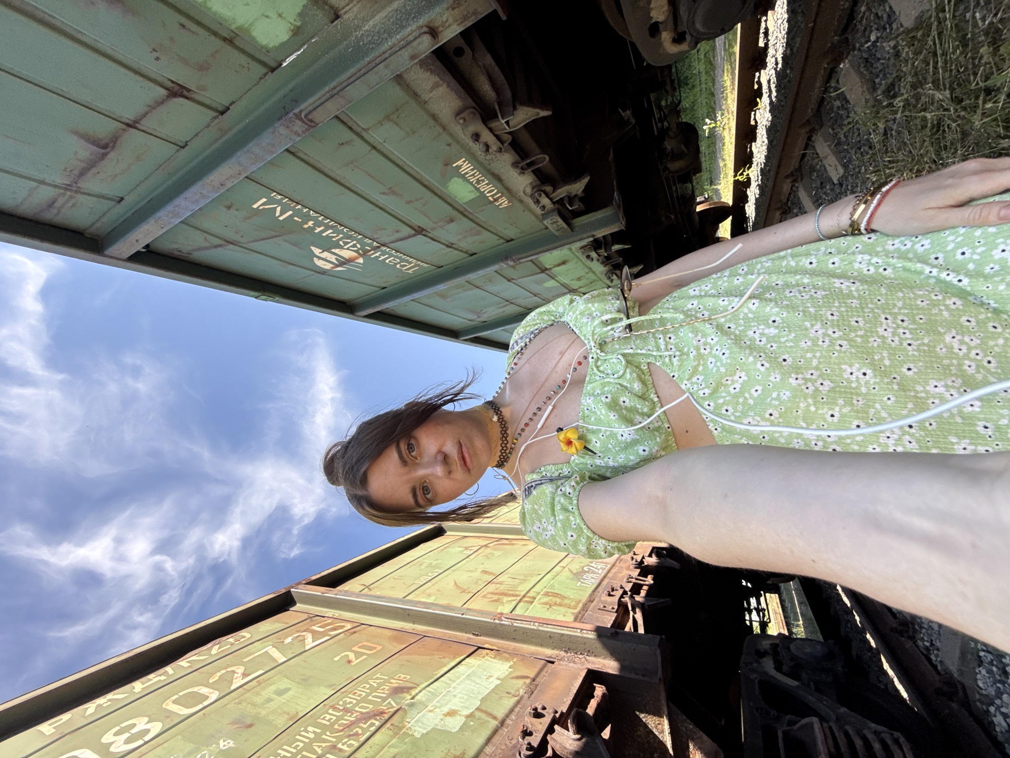 A young woman with brown hair styled in a bun takes a selfie outdoors. She is standing between two large, green, weathered train cars on a railway track. She is wearing a light green, floral-print crop top with cutouts and a yellow flower accessory. She has multiple necklaces and bracelets on and appears to have earbuds in her ears. The sky above is blue with wispy clouds. The train cars have visible markings and numbers.
