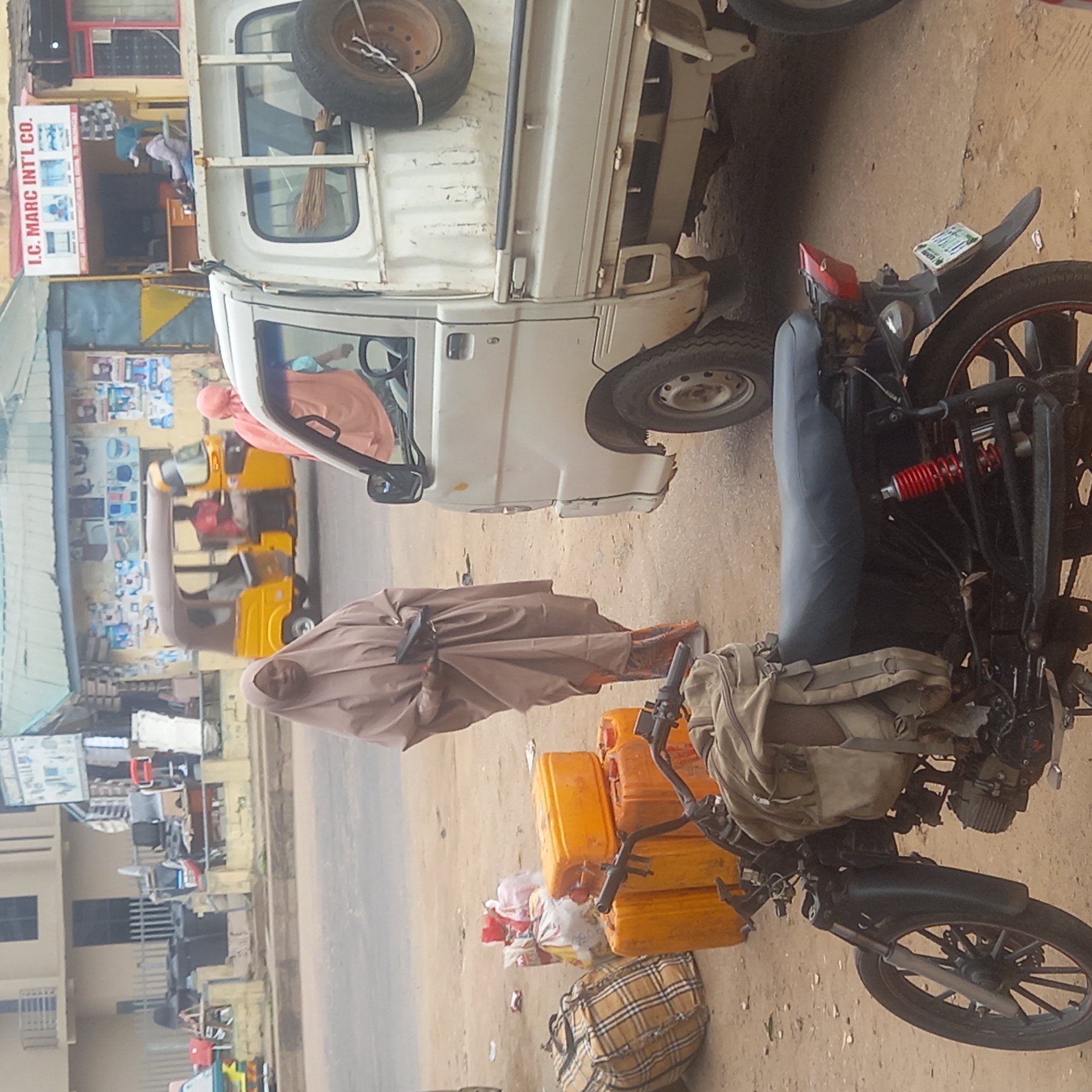 In Dadi Riba, Nigeria, a person in a brown garment stands next to a black motorcycle laden with yellow jerrycans and a checkered bag. The motorcycle is parked on a dirt road next to a small white truck. To the left of the person, there's a yellow auto-rickshaw, and a building with a sign that reads "I.C. MARC INT'L CO." is visible in the background, along with other buildings and urban elements.