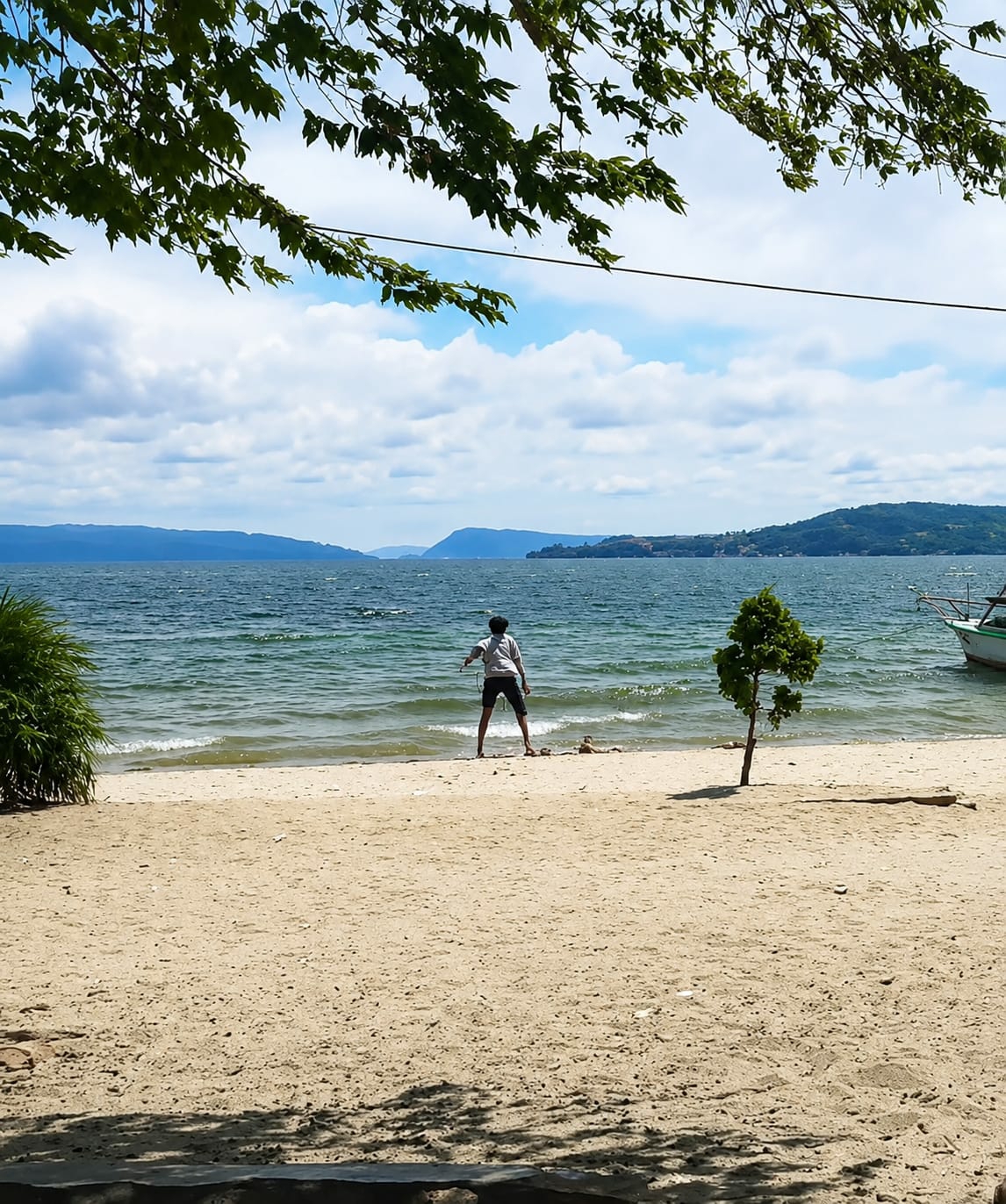 A person stands on a sandy beach facing a body of water. The person is wearing a light-colored shirt and dark shorts, with their arms slightly outstretched. The beach is composed of light-colored sand and some scattered debris. To the right of the person, a small, conical evergreen tree grows. The water is a deep blue-green with visible waves, and in the distance, hills or mountains form the horizon under a cloudy blue sky. A boat with a white hull and green trim is moored on the right side of the water. Overhead, dark green leaves from a tree frame the top of the image. The overall scene suggests a natural, possibly recreational, setting at the edge of a lake or sea. The location is Lumban Bul Bul, Indonesia.