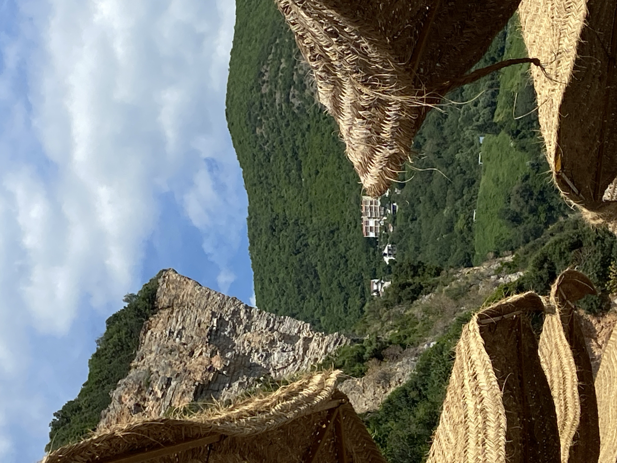 The image presents a scenic view, likely captured in the vicinity of Prijevor, Montenegro, given the distinctive mountainous terrain and coastal vegetation. The viewpoint is from beneath a straw-thatched structure, possibly a beach umbrella or palapa. The foreground is dominated by the textures of the straw, partially obscuring the panorama.
In the middle ground, a steep, green mountainside is visible. The vegetation appears dense, suggesting a lush, Mediterranean environment. Higher up, a cluster of white buildings is observed, perhaps indicating a small settlement or residential area clinging to the hillside. To the left, a prominent rocky outcrop with sparse vegetation contrasts with the green slopes. A blue sky dotted with scattered clouds is visible in the background. The absence of people is noted.