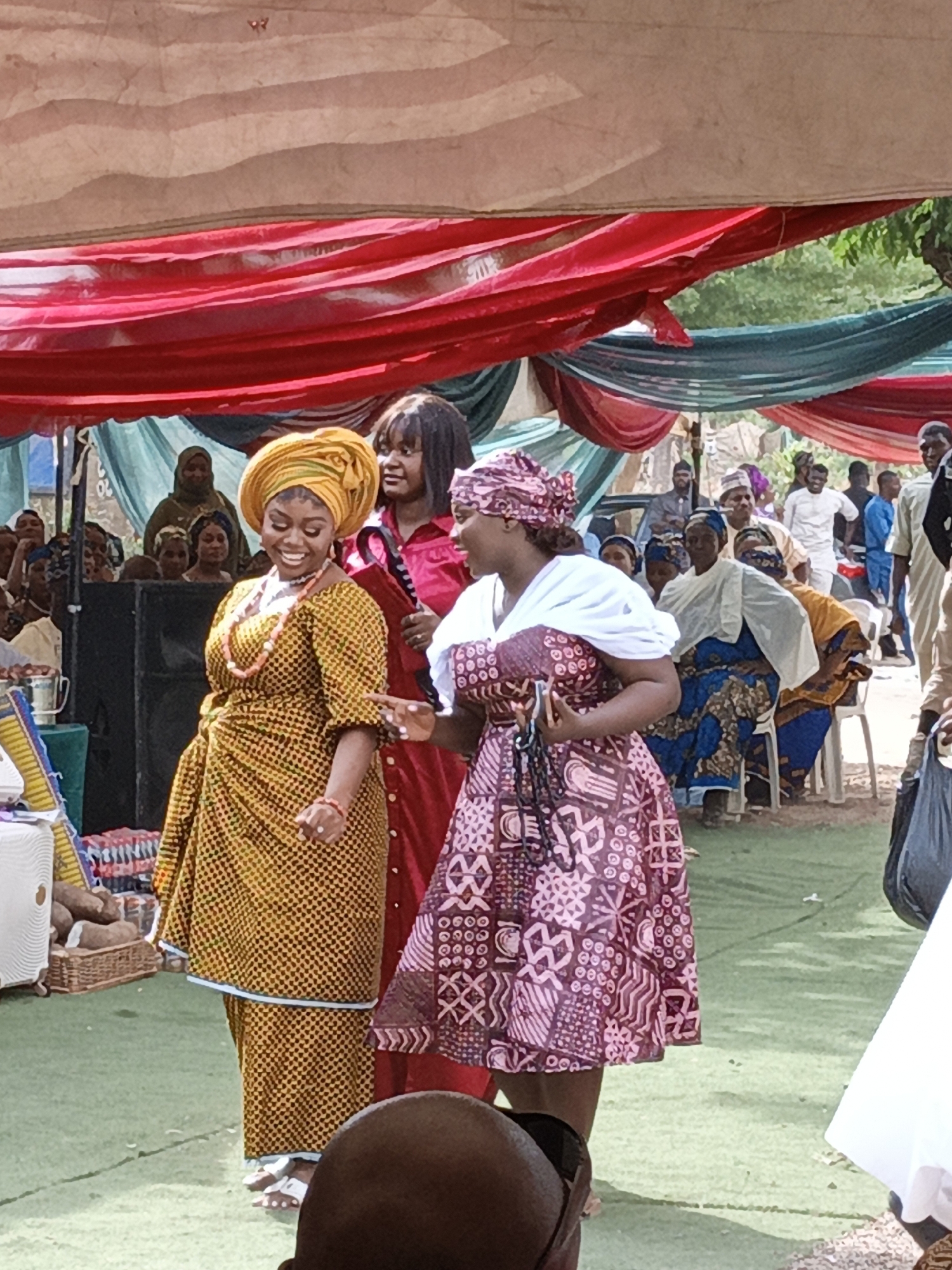This image captures a vibrant outdoor celebration, likely in Sabon Gari West, Nigeria, under a large canopy draped with red and teal fabrics. The ground is covered with a green surface, possibly artificial turf. The scene is brightly lit, suggesting daytime.
In the foreground, three women are prominently featured, all dressed in traditional West African attire. On the left, a woman beams with a wide smile, wearing a bright yellow headwrap (gele) and a matching yellow Ankara print dress with small black patterns. She accessorizes with a coral beaded necklace and bracelet and wears white sandals. She appears to be dancing or moving animatedly. Behind her, a woman with dark, shoulder-length hair wears a long-sleeved red dress and also smiles, looking towards the first woman. To the right, another woman wears a patterned headscarf and a brownish-pink and white patterned dress, possibly with white sleeves. She is facing the yellow-dressed woman, gesturing with her hand, suggesting an interaction or conversation. She holds a small, dark object in her hand.
In the background, numerous other attendees are visible, both standing and seated on white plastic chairs. Many are also dressed in patterned traditional clothing and headwraps, observing the foreground activity. A large, dark speaker is positioned on the left side of the scene, indicating music or amplified sound. Nearby, a woven basket contains what appears to be yams or similar produce, alongside a white cooler or stand. The overall atmosphere appears joyous and festive. No discernible text is visible in the image.