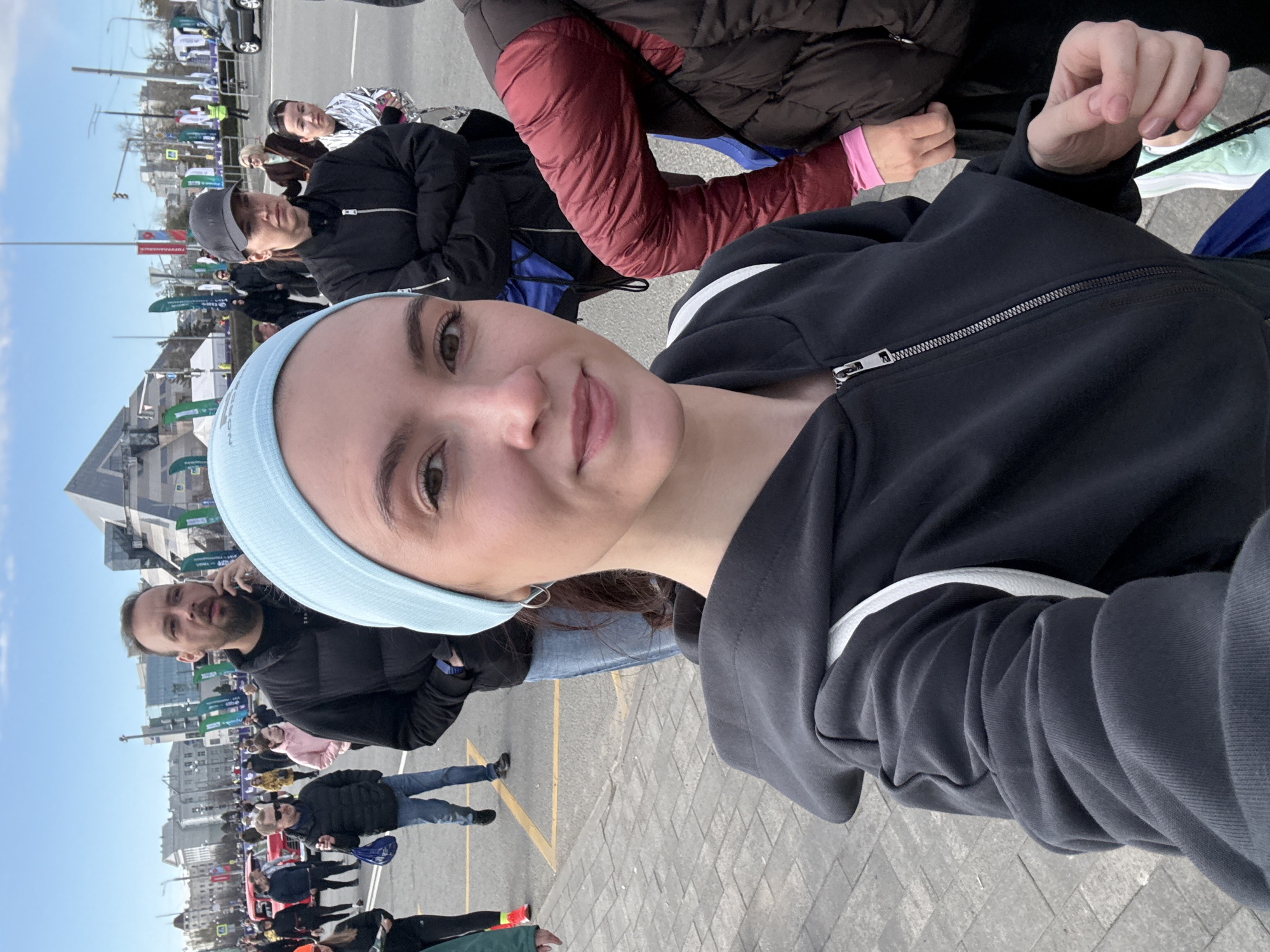A woman wearing a light blue headband and a black zip-up hoodie is taking a selfie. She is standing in a crowd of people on a paved area marked with yellow lines. In the background, there are buildings, flags, and signs. A man behind her appears to be on the phone. The scene suggests an outdoor event, possibly a race or gathering, located in Kazan, Russia.