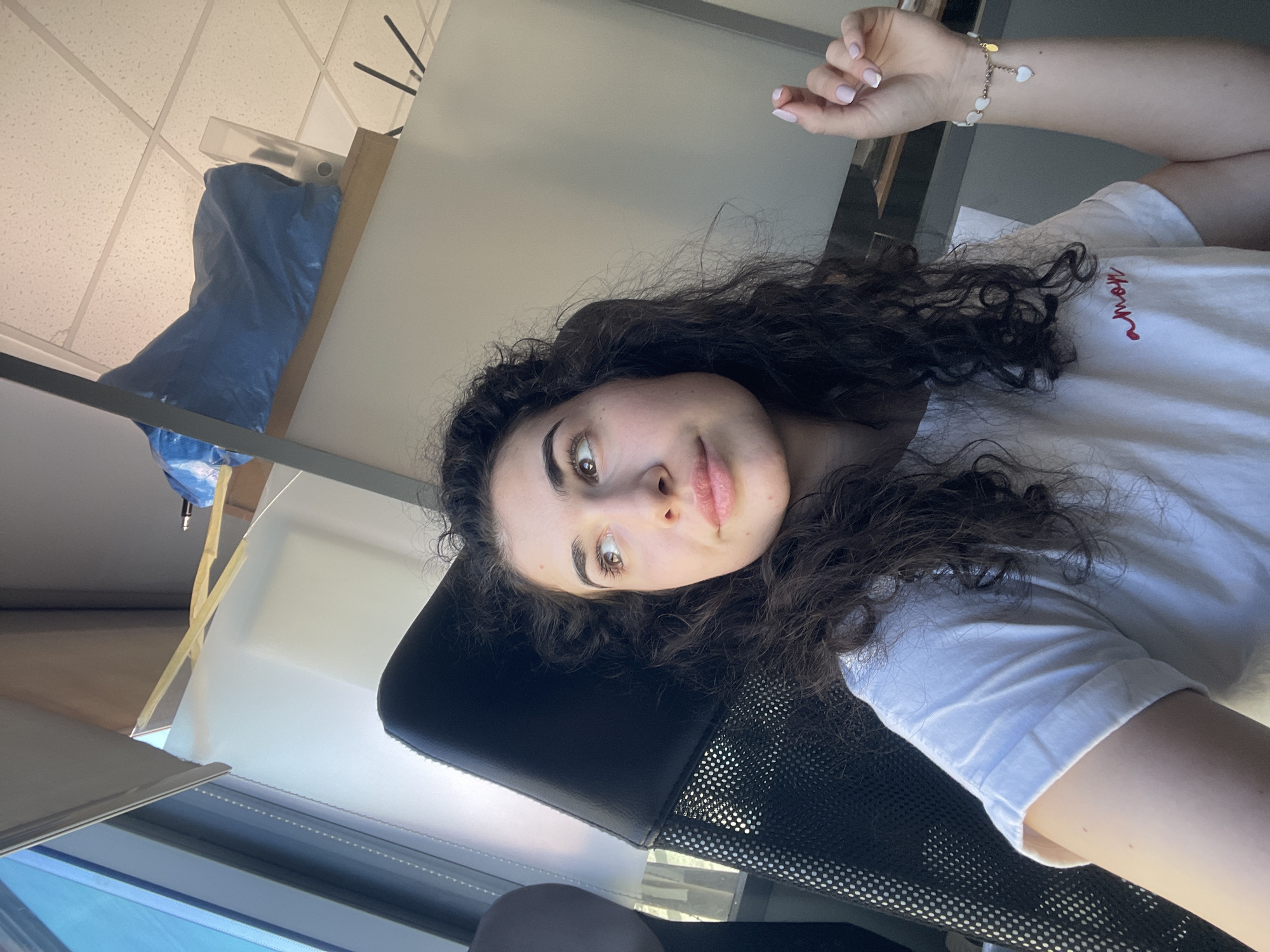 A medium-range shot features a woman in a white t-shirt, seated in a black office chair, looking towards the camera. Her arm is visible, adorned with a beaded bracelet. A window is visible in the lower-left corner. A blue bag is situated above her. The location is likely an office space, with a drop ceiling. The city of Sofia, Bulgaria is not discernible from the given visual information.