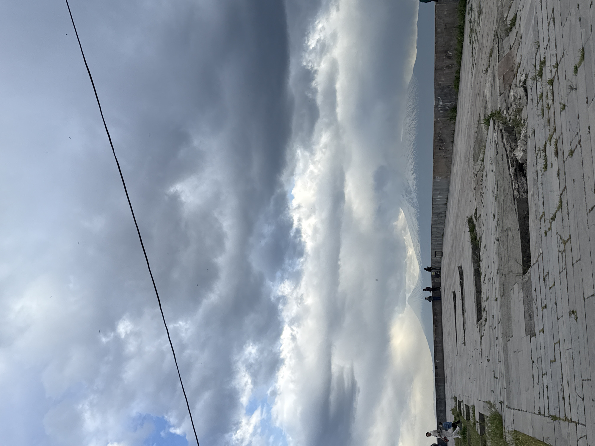 The image depicts a partly cloudy sky over Yerevan, Armenia, with a view of Mount Ararat in the background. A dark electrical wire diagonally crosses the upper left of the frame. To the right is a light gray, stone or concrete structure with several rectangular openings, possibly part of a building or monument. People can be seen standing or sitting near the structure, some of whom appear to be looking at Mount Ararat. The foreground includes grass and vegetation growing near the stone structure, suggesting it may be located in a park or public space.