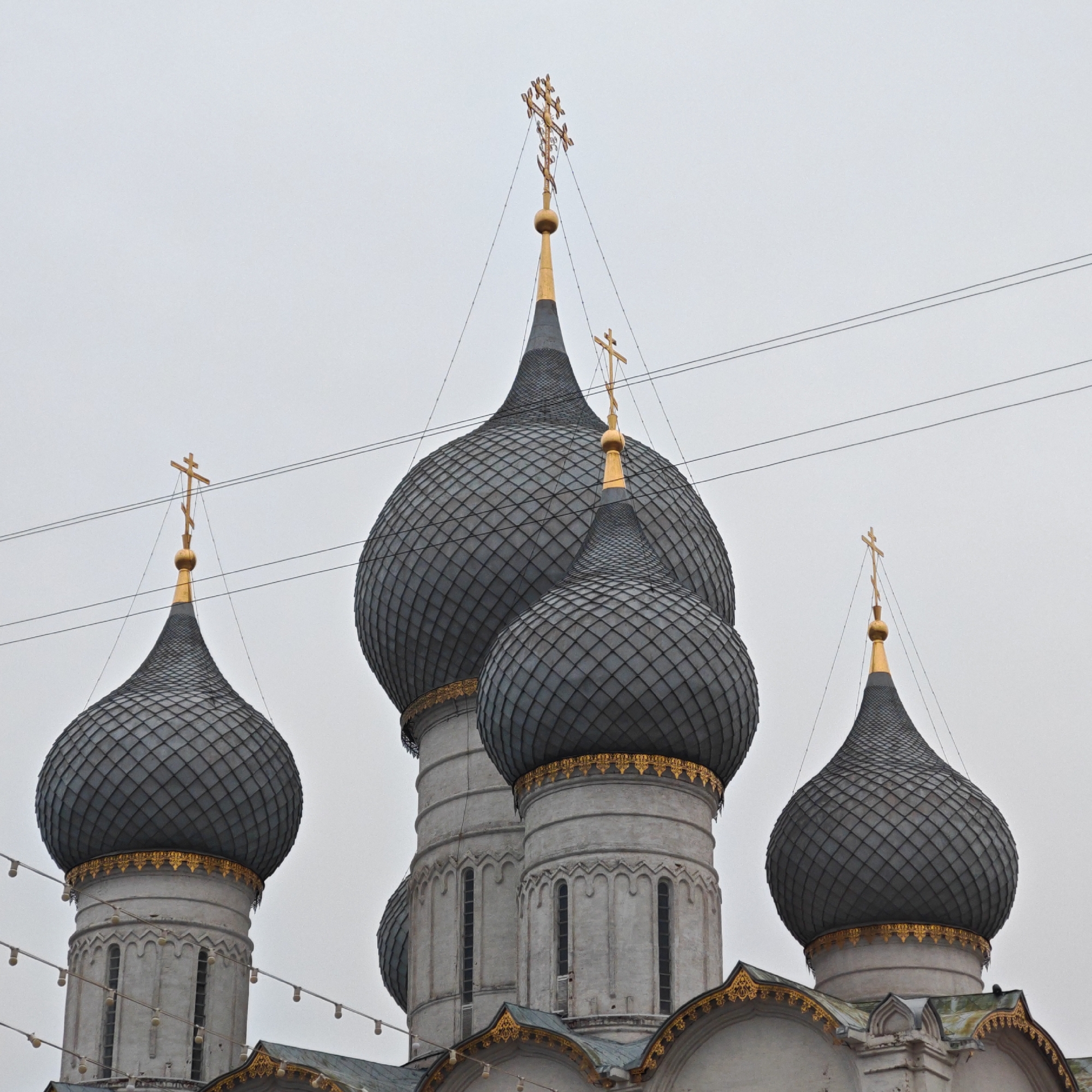 The image depicts a partial view of a Russian Orthodox church, identified as being located in Rostov, Russia. The primary focus is on four onion-shaped domes covered in dark gray, scale-like patterns. Each dome is topped with a golden finial and a Russian Orthodox cross. The base of each dome is adorned with gold ornamentation. The towers supporting the domes are white and have vertical slit windows. Power lines are visible against the gray sky, which suggests an overcast day. The architecture has detailed golden roof trim at the base of some domes.
