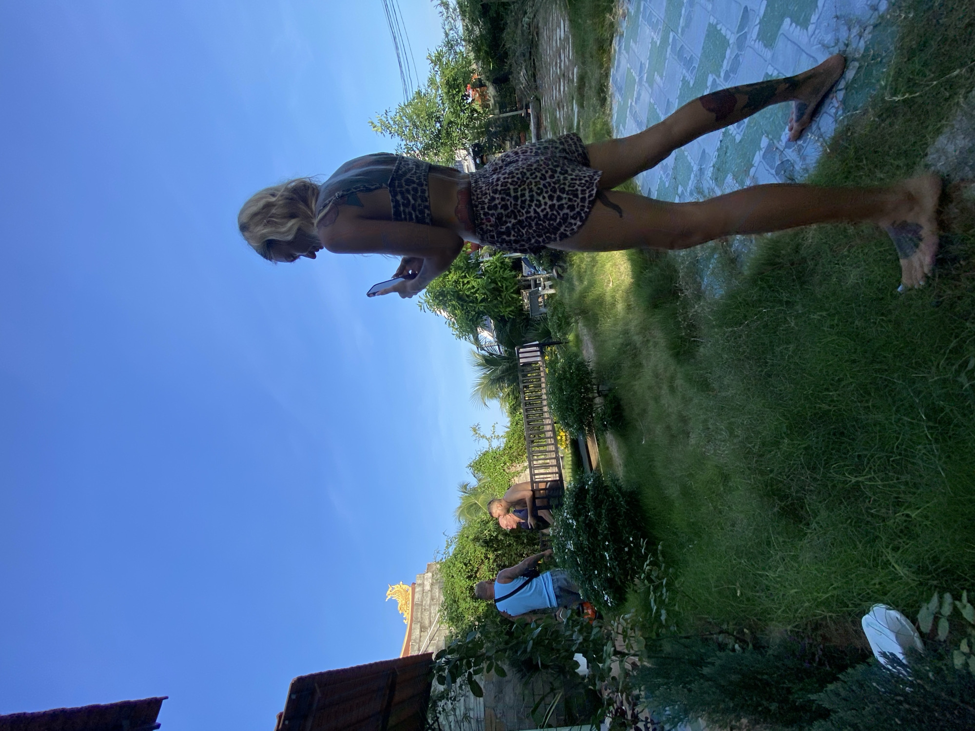 In Mũi Né, Vietnam, a woman with blonde hair, wearing a leopard print bikini and matching shorts, stands in a grassy yard. She is looking down at her phone, which she holds in her right hand. The woman has tattoos on her back, legs, and feet. In the background, two men are talking near a wooden railing. One man wears a blue tank top and denim shorts, while the other is shirtless and wears dark shorts. Behind them, a building with a golden ornate roof is visible. The environment appears to be a tropical outdoor space with lush green grass, various plants, and palm trees. The sky is clear and blue. The ground is a tiled patio.