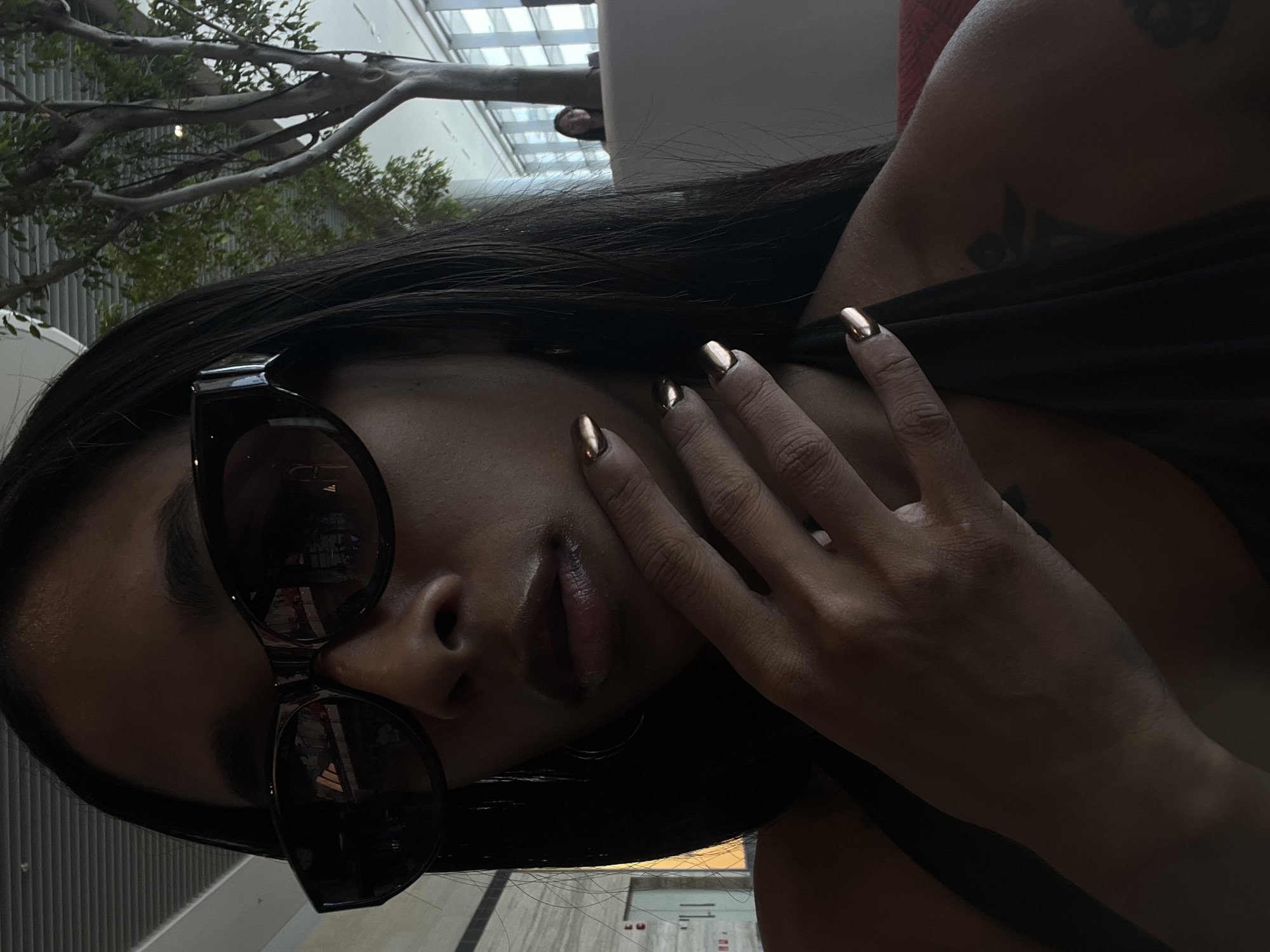 A woman with long dark hair and large dark sunglasses is shown in a close-up, selfie-style shot. She is wearing a black sleeveless top and has her right hand, with gold-colored nails, resting near her chin and neck. A tattoo is visible on her left shoulder. The background suggests an indoor, modern setting with large windows, a tall indoor tree with green foliage, and architectural elements like a metal ceiling grid and what appears to be a light-colored partition. In the distance, another person is visible through the windows. The overall lighting is dim, with the woman's face and hand illuminated. Based on the presence of indoor trees and modern architecture, the location is likely an atrium or a well-designed commercial space within Singapore.