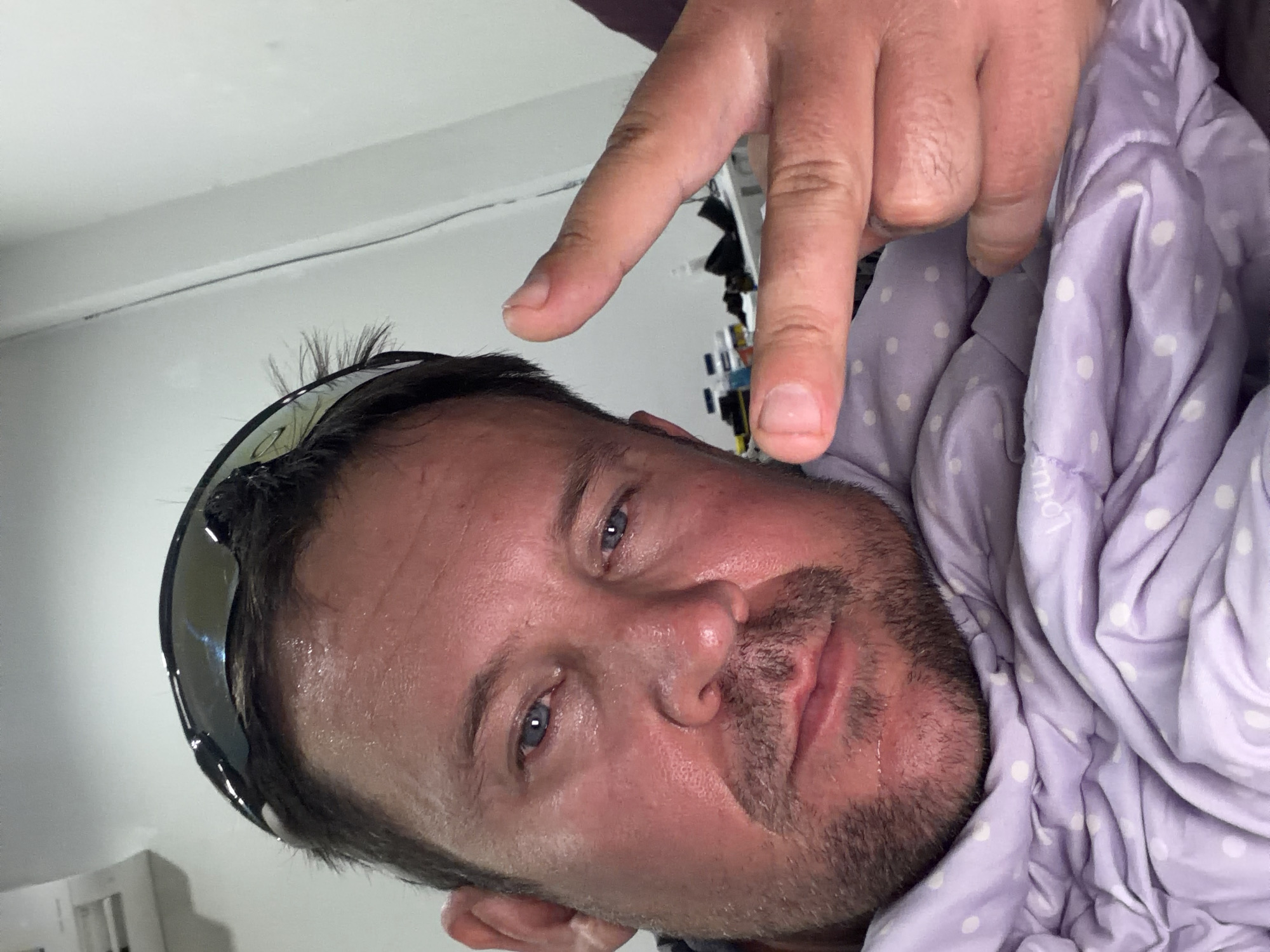 A man, positioned in the foreground, is lying down, covered by a light purple polka-dot blanket. He is wearing black sunglasses on his head and appears to be sweating. His right hand is raised, with his index and middle fingers extended in a peace sign gesture. The background shows a white wall with some items on a shelf, including bottles and what appears to be a white air conditioning unit. The scene is indoors, likely in a room within Baan Mae Nam, Thailand.