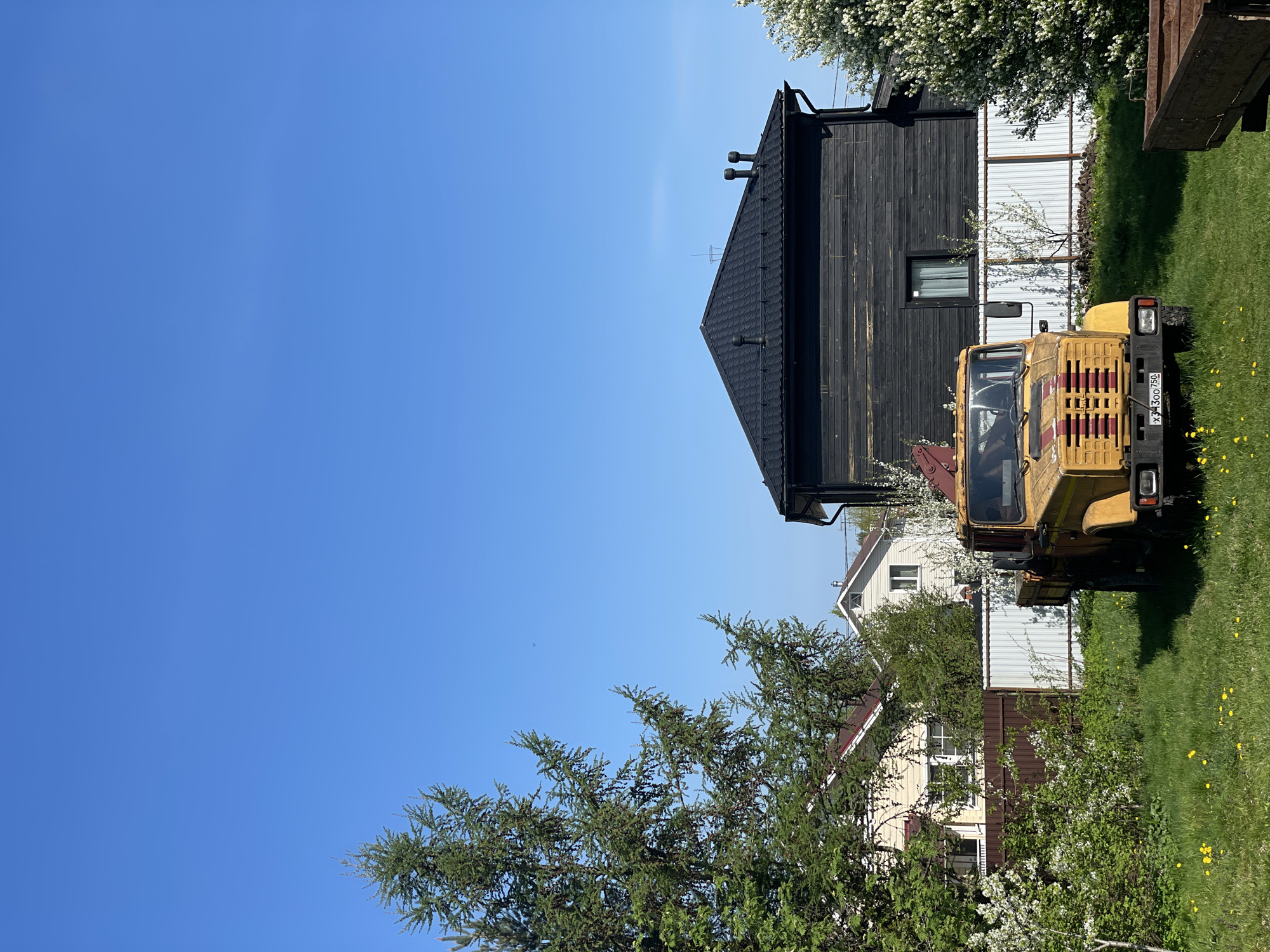 A yellow and red all-terrain vehicle is parked on a grassy lawn in Sestroretsk, Russia. Behind the vehicle is a black wooden building with a white fence, partially obscured by blossoming trees. In the background, other houses with light-colored siding and red or brown roofs are visible. The sky is clear blue. A coniferous tree is in the foreground.