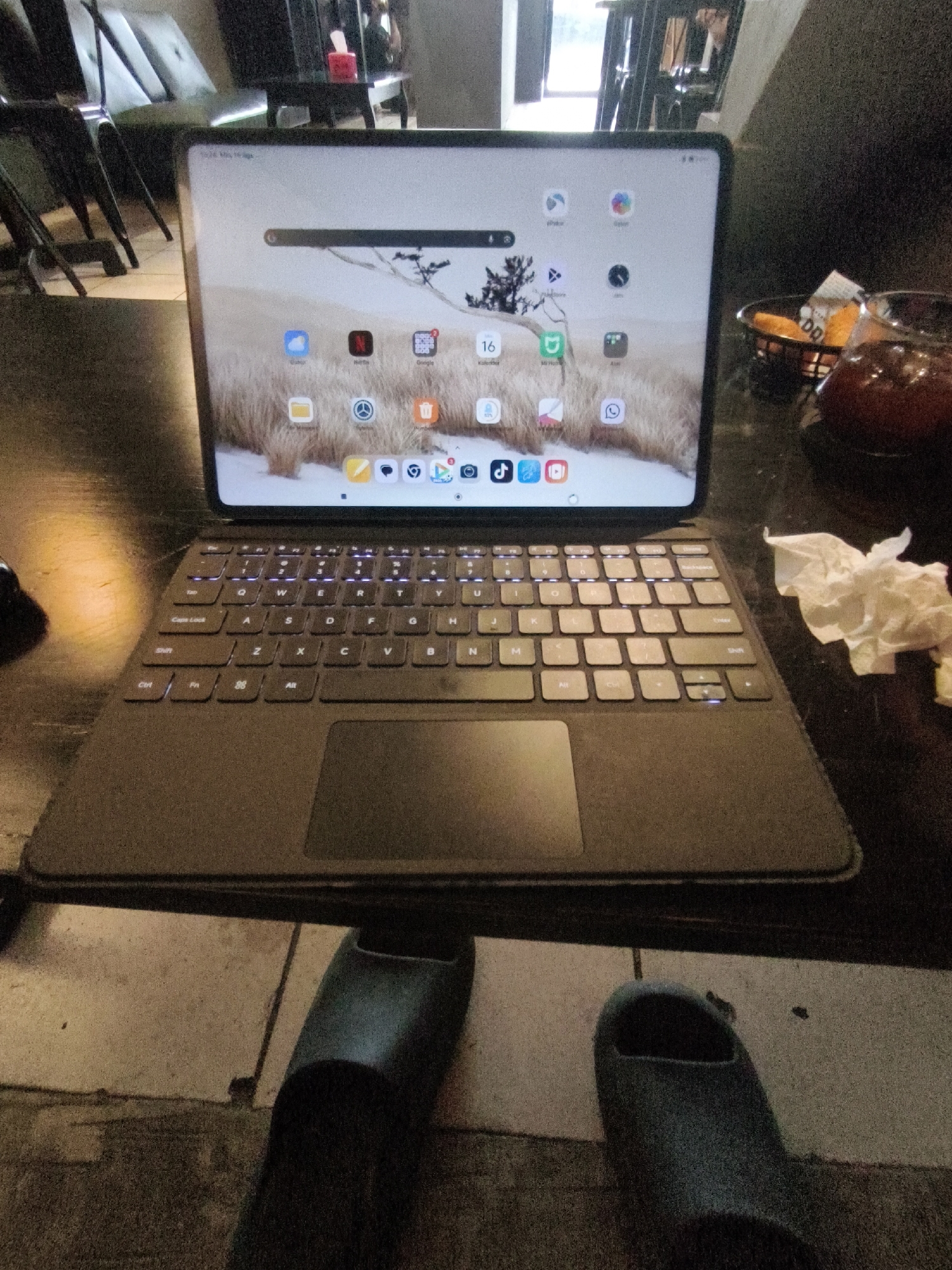The image shows a tablet with a keyboard attachment resting on a dark wooden table. The tablet's screen displays a wallpaper of a snowy landscape with trees and a search bar. Several app icons are visible on the screen, including ones for "Cloud," "Netflix," "Google," and "Calendar." The keyboard is illuminated with backlighting. To the right of the tablet, there is a small basket containing what appear to be snacks and a glass of dark liquid. A crumpled white tissue is also visible near the glass. In the foreground, a pair of dark blue slippers are placed on a tiled floor. In the background, a blurred view of a seating area with chairs and a couch is visible. The overall setting appears to be an indoor public space.