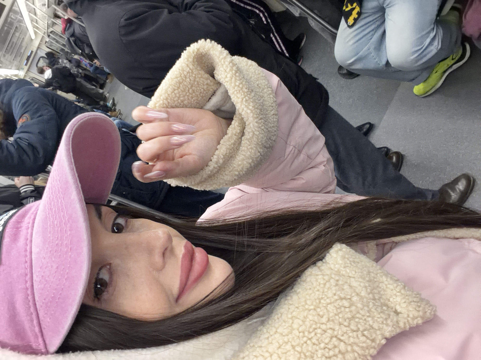 The image shows a young woman with brown hair and brown eyes wearing a pink baseball cap and a pink jacket with a cream-colored sherpa lining. She is in what appears to be a public transportation setting, possibly a train, judging by the presence of other passengers in the background. Some of these passengers are wearing winter clothing and are seated. There is a person wearing sneakers and light blue jeans in the background. The location could be in Saint Petersburg, Russia, given the typical weather in the city and the overall style of clothing worn by the people in the image.