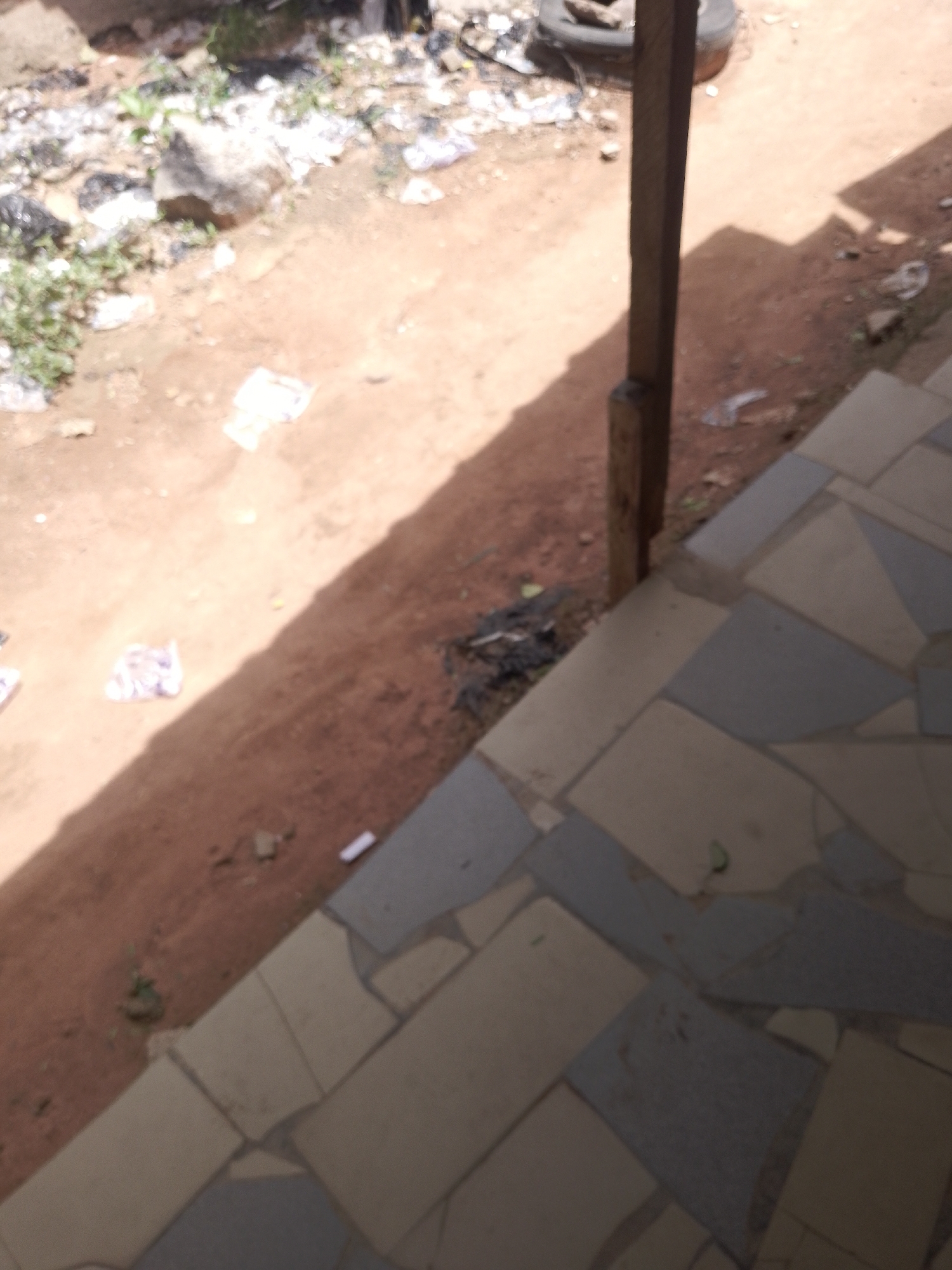 An outdoor, high-angle, slightly blurry photograph shows a dirt path with scattered debris, a wooden post, and a tiled walkway in the foreground. The dirt path occupies the left and upper portions of the image, with various items strewn across it, including rocks, dry leaves, and what appear to be pieces of white plastic or paper. A large, dark, circular object, possibly a tire, is visible in the upper left quadrant. A thick, weathered wooden post stands vertically in the middle right of the frame, casting a prominent shadow that stretches across the dirt path. To the right of the post and in the lower half of the image, a tiled walkway is visible, made up of irregularly shaped tiles in shades of beige and blue-gray, laid in a mosaic pattern. The ground appears to be uneven, with some areas of exposed dirt and small patches of vegetation. The overall impression is of a somewhat unkempt outdoor area.
