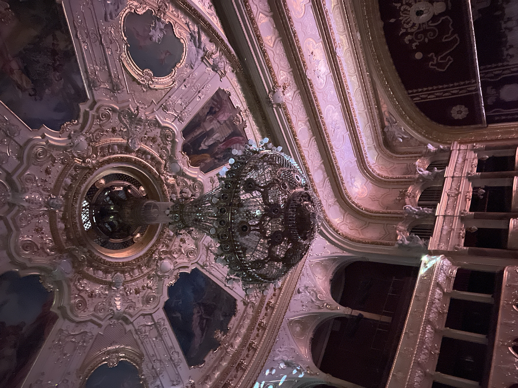 The image captures a detailed view of the ceiling and a chandelier in what appears to be an ornate theater or opera house, possibly the Odesa Opera and Ballet Theater in Odesa, Ukraine. The ceiling features intricate plasterwork, paintings within decorative frames, and gold detailing. A large, ornate chandelier hangs in the center, its design featuring multiple tiers of lights and intricate metalwork. To the right, there are architectural elements suggesting theater boxes or balconies, with ornamental carvings and dark arched openings. The scene is richly decorated, indicative of a historical and culturally significant venue.
