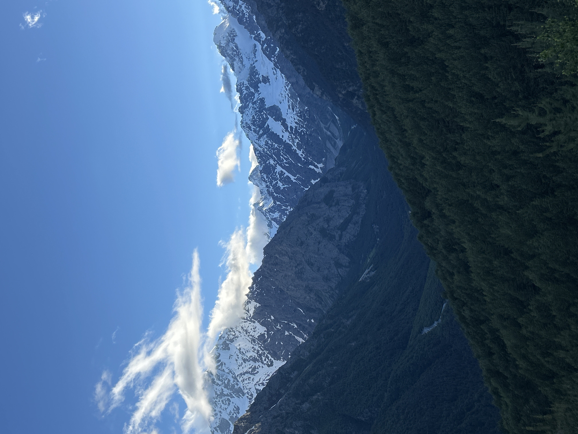 The image shows a majestic mountain landscape with snow-capped peaks against a clear blue sky. The foreground is dominated by a dense forest of dark green trees covering steep slopes. In the distance, rugged mountains rise, their upper reaches dusted with snow, catching the sunlight. Wispy clouds drift across the sky. The scene appears to be a natural, undeveloped area, likely in a mountainous region. Without further information, it is not possible to determine if this image is of Abaitikau, Russia, or to provide any details about people, actions, or interactions.