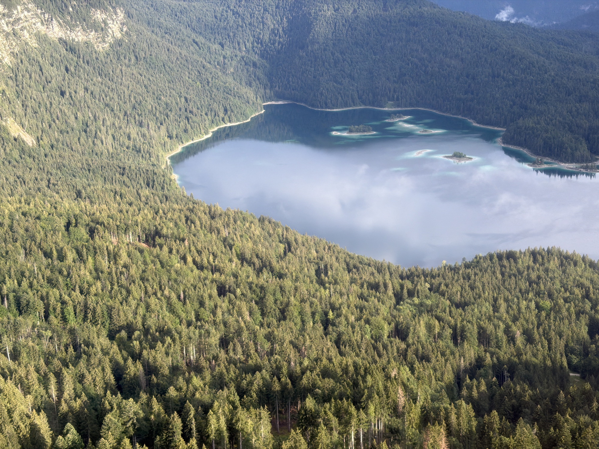 An aerial, eye-level, wide-angle shot captures a vast, dense evergreen forest on a steep mountainside sloping down to a calm, reflective lake. The forest is a mix of dark green and lighter green trees, with patches of sunlight breaking through, creating a dappled effect. The lake, a deep blue-green, occupies the center and right portion of the image, its surface broken by several small, tree-covered islands. The water is calm, reflecting the surrounding trees and the sky above, with wispy white clouds visible. The shore of the lake is lined with the dense forest, with a few rocky outcrops visible on the far side. The scene is serene and natural, with no signs of human activity. The image is likely taken in the vicinity of the Eibsee near Grainau, Germany, a well-known glacial lake at the base of the Zugspitze mountain.
