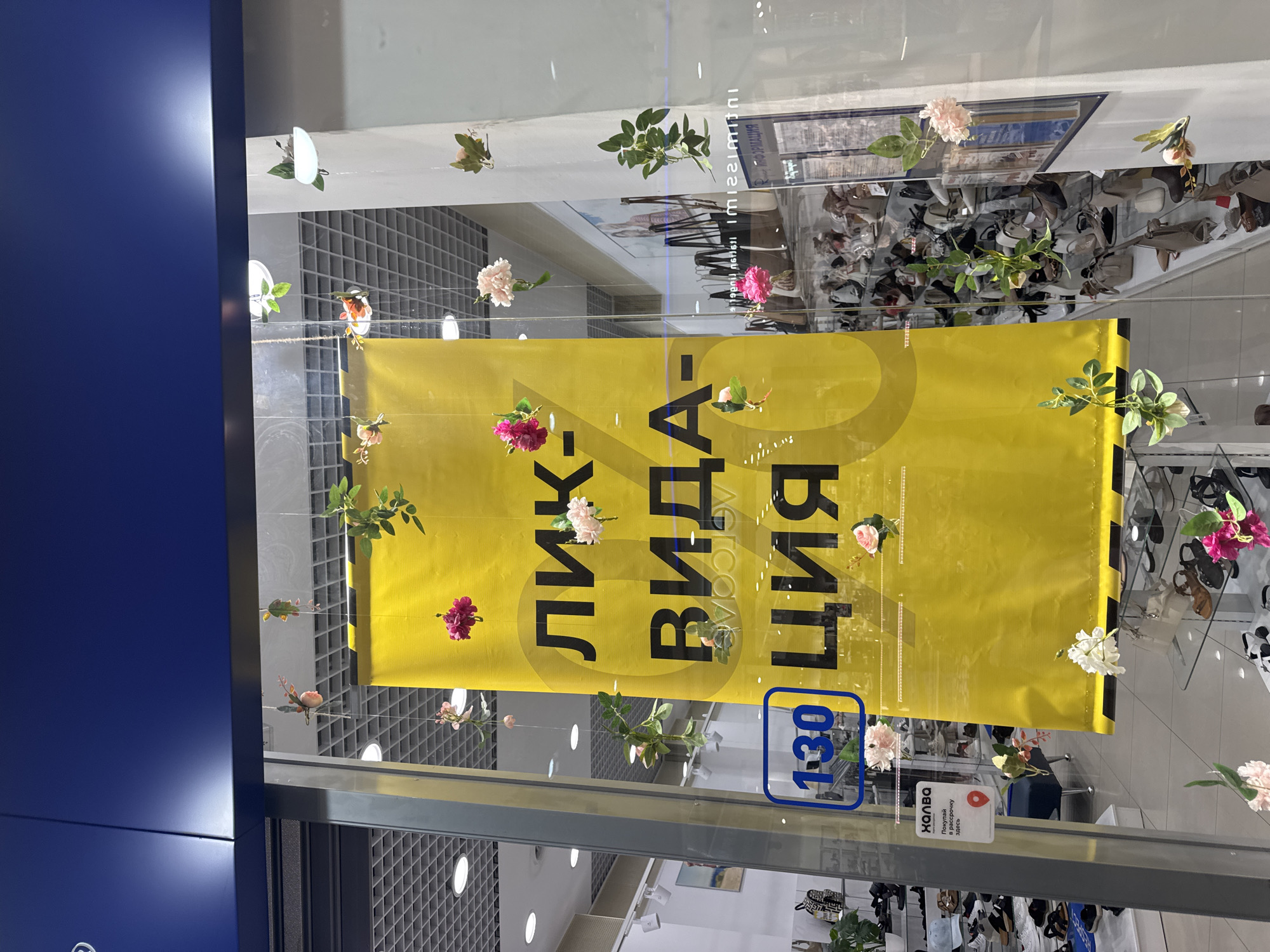 Here is a description of the image.
The image shows a storefront window display. A large yellow banner with Russian text "ЛИКВИДАЦИЯ" (liquidation) is prominently displayed. The banner has a large circular design element behind the text. Artificial flowers and leaves are suspended in front of the banner and near the window edges. Through the window, shoes are visible on shelves inside the store. A small sign with the word "ХАЛВА" is visible on the lower portion of the glass. A blue sign with white numbers "1-30" is also on the glass. The location and city are unknown.