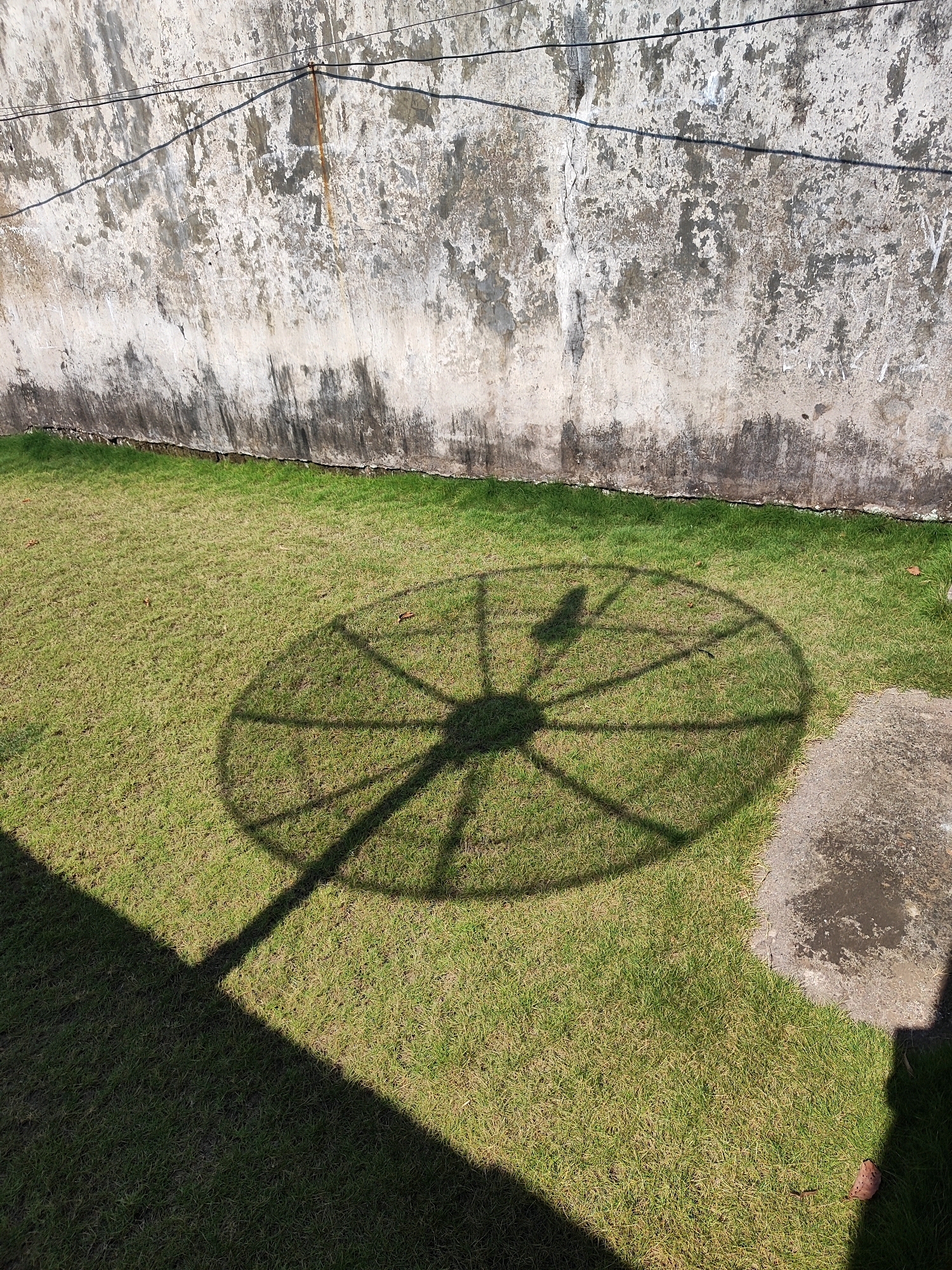 A satellite dish's shadow is cast on a patch of green grass in front of a weathered, textured wall. The wall, likely a building exterior, displays signs of age with patches of peeling paint and dark streaks, possibly from water damage or dirt. Two thin wires stretch across the top of the wall, casting faint shadows of their own. The grass is neatly trimmed and appears to be a well-maintained lawn. To the right of the grass, a concrete slab with a rough, uneven surface is visible, also partly in shadow. The scene is illuminated by bright sunlight, creating a strong contrast between light and shadow. The image appears to have been taken outdoors, likely in a residential or communal area within the city of Kotakulon, Indonesia. No people are visible in the frame.