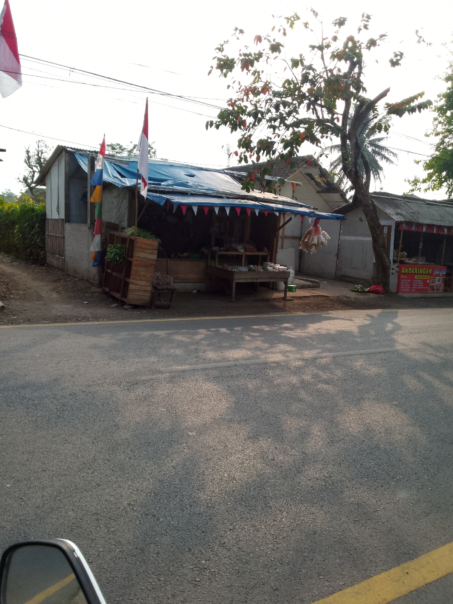 The image depicts a street scene in Haurwangi, Indonesia, featuring a small roadside stall. The stall has a blue tarp roof adorned with red and white triangular pennants. Various items are displayed, including what appears to be vegetables in a wooden crate on the left, and hanging bags of goods above a table. To the right of the stall, a red sign with white lettering, likely a menu, is visible. Indonesian flags are displayed, one prominently on a pole to the right of the stall and another, partially visible, on the left. The stall is situated on the side of a paved road, with a yellow line marking the edge of the road. The environment is characterized by a tree with green and red leaves to the right of the stall, and a building with a corrugated metal roof behind it. The sky is overcast. The foreground of the image shows the asphalt road and a portion of a motorcycle's side mirror on the bottom left, suggesting the photo was taken from a moving vehicle. Shadows of trees and possibly a person are cast on the road.