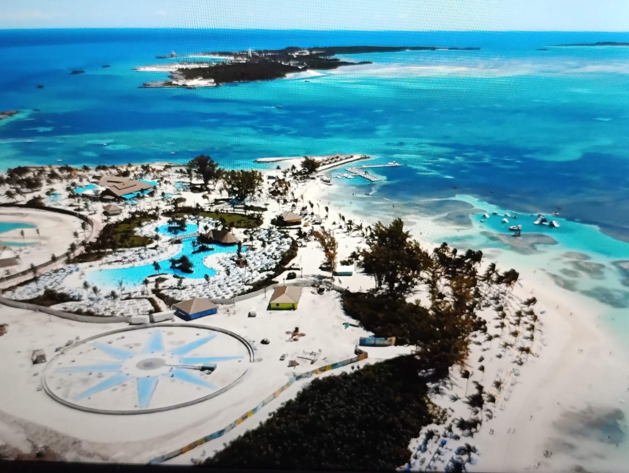 This is an aerial photograph of a tropical island resort. The scene features multiple swimming pools, thatched-roof buildings, and a large, star-shaped structure on the sandy ground. The island is surrounded by clear turquoise water and a sandy beach lined with palm trees. Several boats are visible in the water near a pier. In the background, another island with vegetation and buildings can be seen in the distance. The sky is clear and blue. The location is not in the city of Dubăsari town council, Moldova, as this is a tropical island resort.