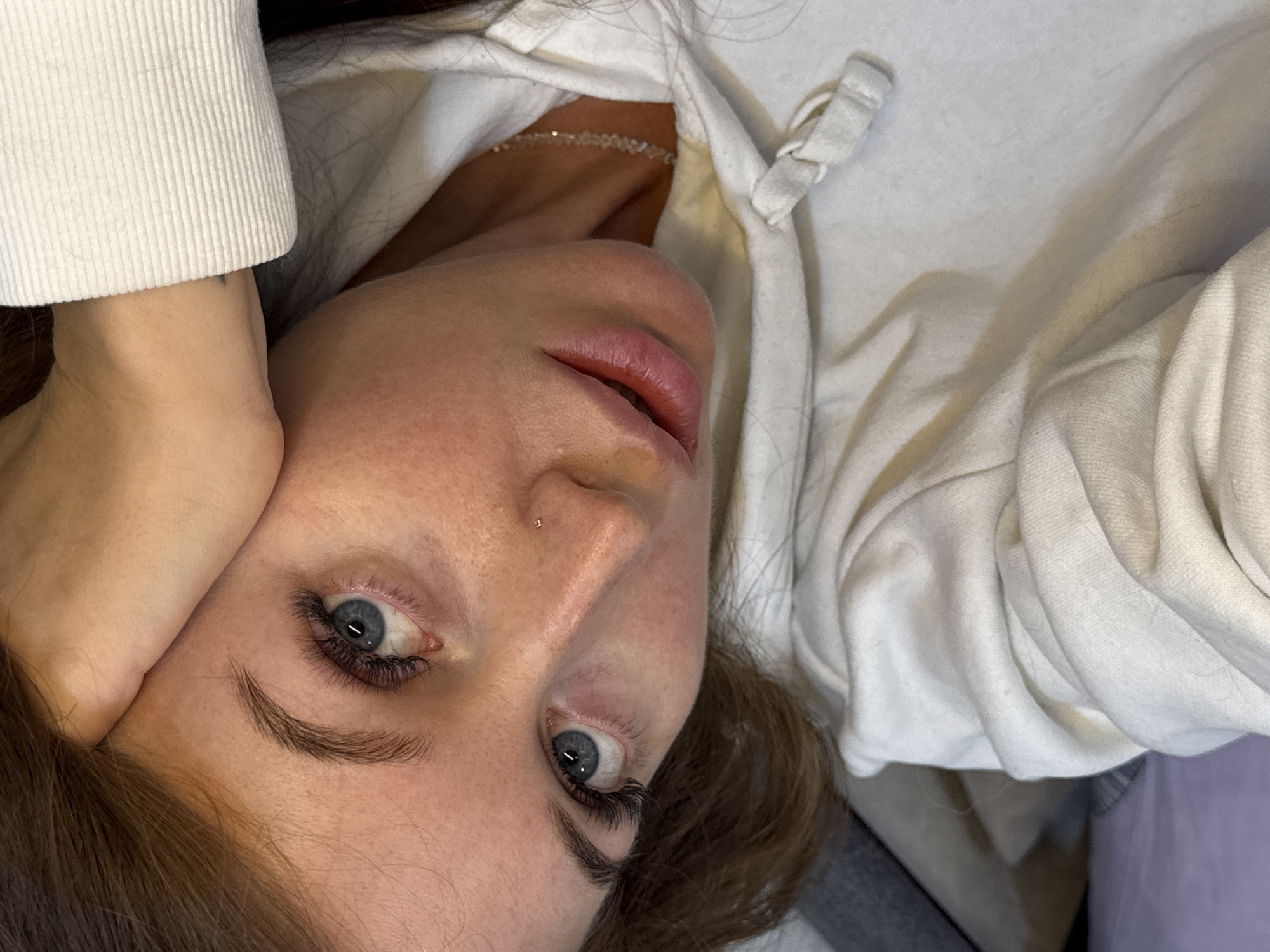 A young woman with blue eyes, brown hair, and a nose piercing is lying down, her head tilted toward the camera. She is wearing a white hoodie and a delicate necklace is visible around her neck. One of her hands is near her face. The background is blurred and indistinct, suggesting an indoor setting. The specific location in Chaikovsky, Russia, is not determinable from the image.
