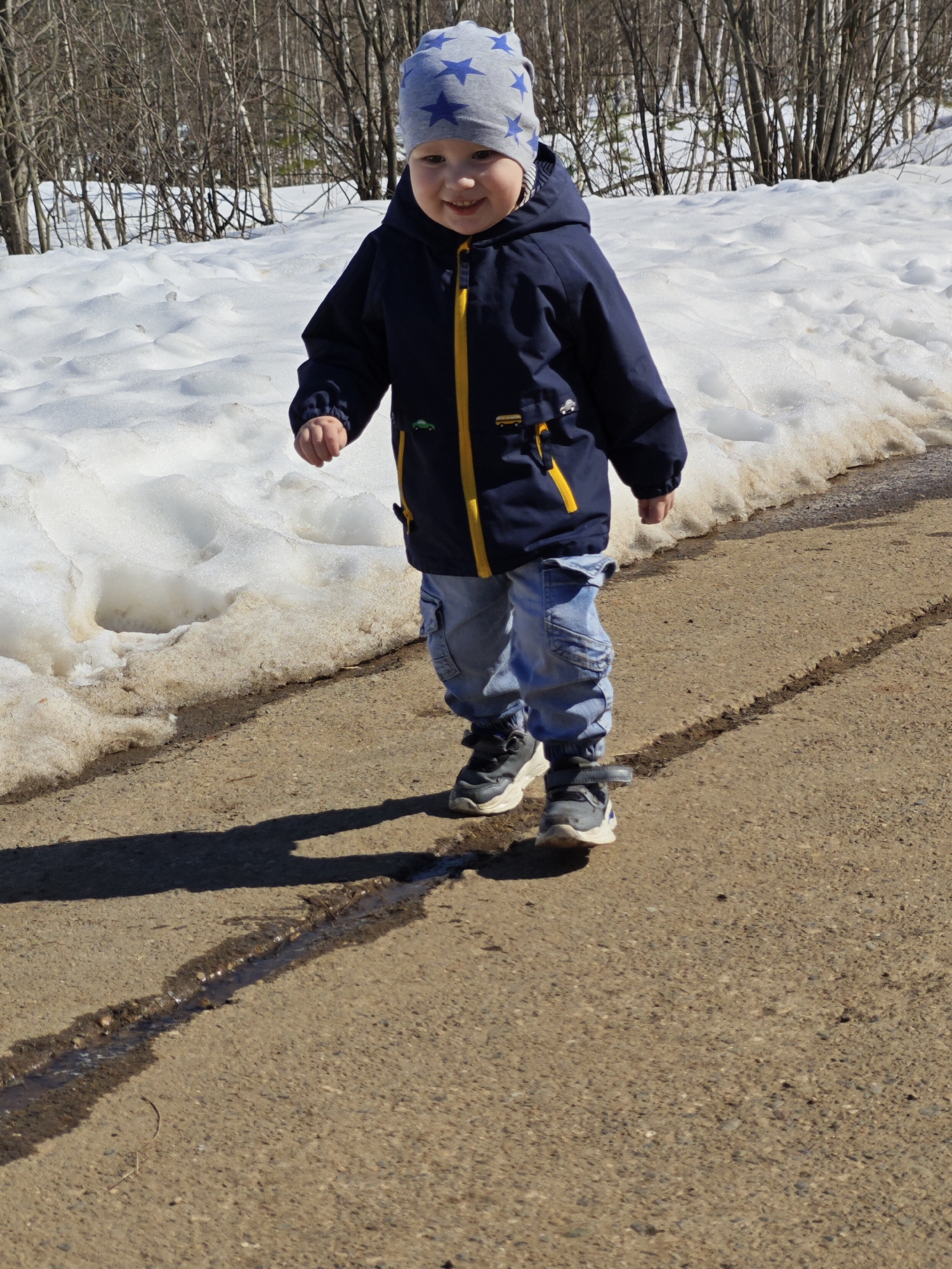 A young child stands on an asphalt path with patches of melting snow nearby. The child wears a blue and grey star-patterned hat, a dark blue jacket with yellow trim, light blue jeans, and dark sneakers. The child is smiling, casting a shadow on the path. The scene is illuminated by bright sunlight. Bare trees are visible in the background. The location is Марковский, Russia.
