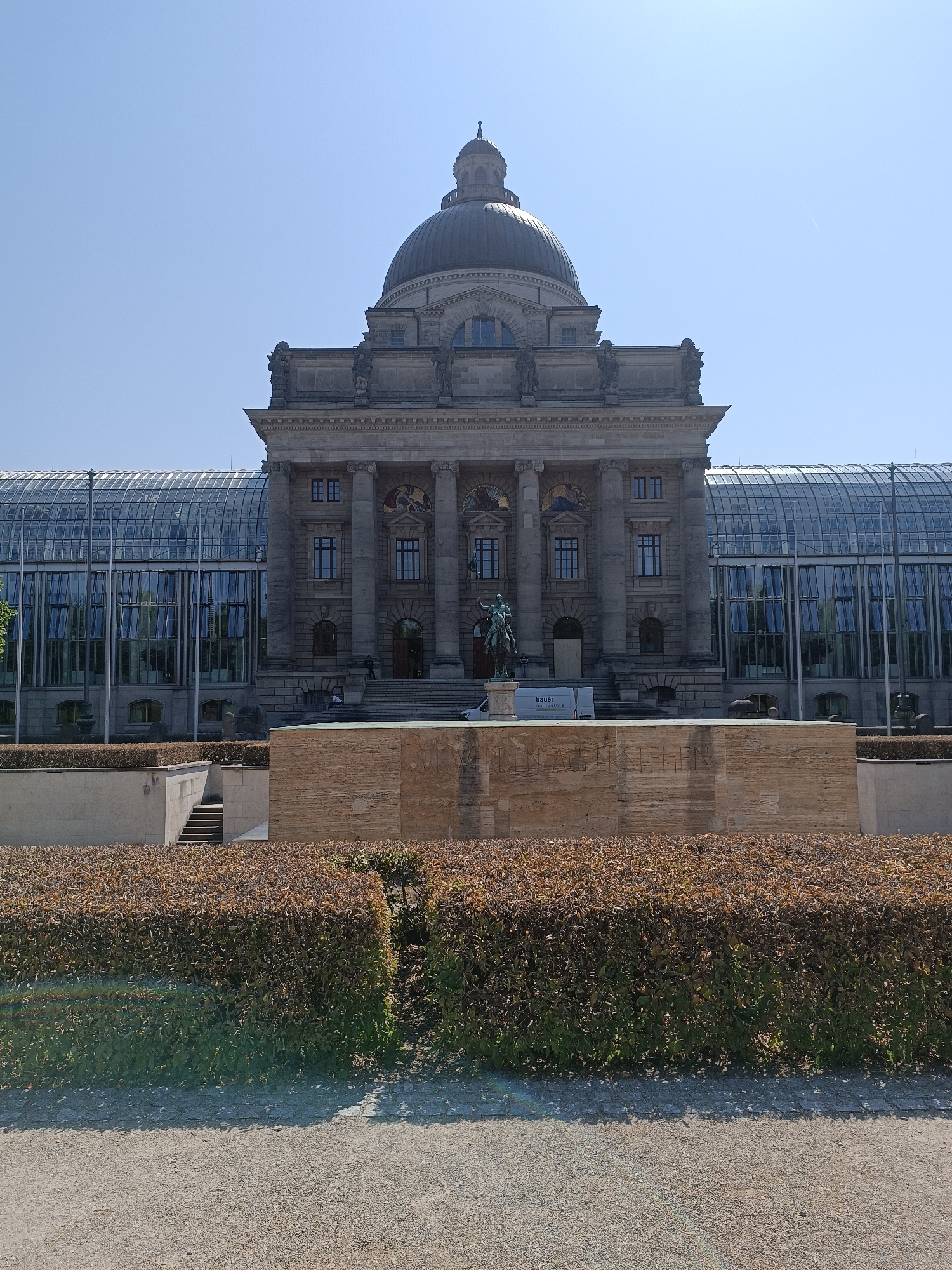 The image depicts the exterior of the Bavarian State Chancellery in Munich, Germany. The central structure features a prominent dome and classical architectural elements with stone columns and facades. To the left and right of the central building are modern glass-paneled extensions that reflect the clear blue sky. In the foreground, a hedgerow with browning leaves partially obscures a tiered stone structure. A bronze equestrian statue stands in front of the central building's entrance stairs. A white van with a logo is parked to the left of the stairs. The ground in the immediate foreground is a gravel pathway.