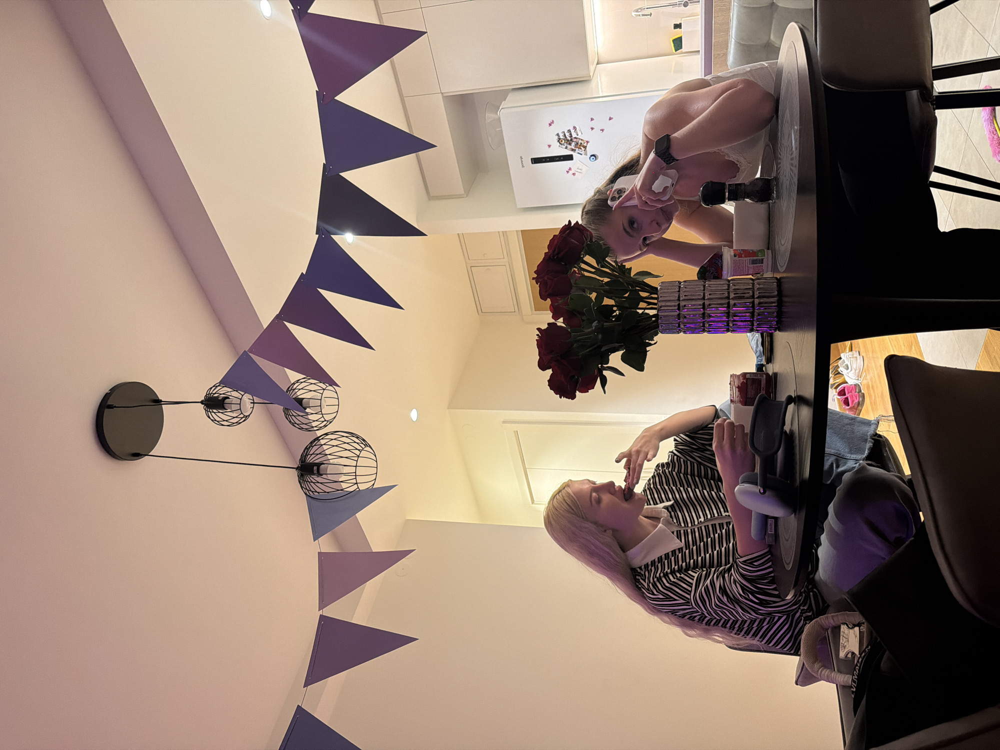 A rotated, brightly lit interior photograph depicts two women at a dining table. Overhead, a triangular purple and blue bunting is hung, alongside a black multi-bulb light fixture. One woman, with blonde hair, sits at the table wearing a black and white striped top and blue jeans; she is holding something to her mouth. On the table, there are wireless headphones. A second woman with long dark hair kneels on the table, facing the camera and holding a white cell phone. A vase of red roses is centrally placed on the table. A white refrigerator is in the background. The precise location within Belgrade, Serbia cannot be determined from the image.