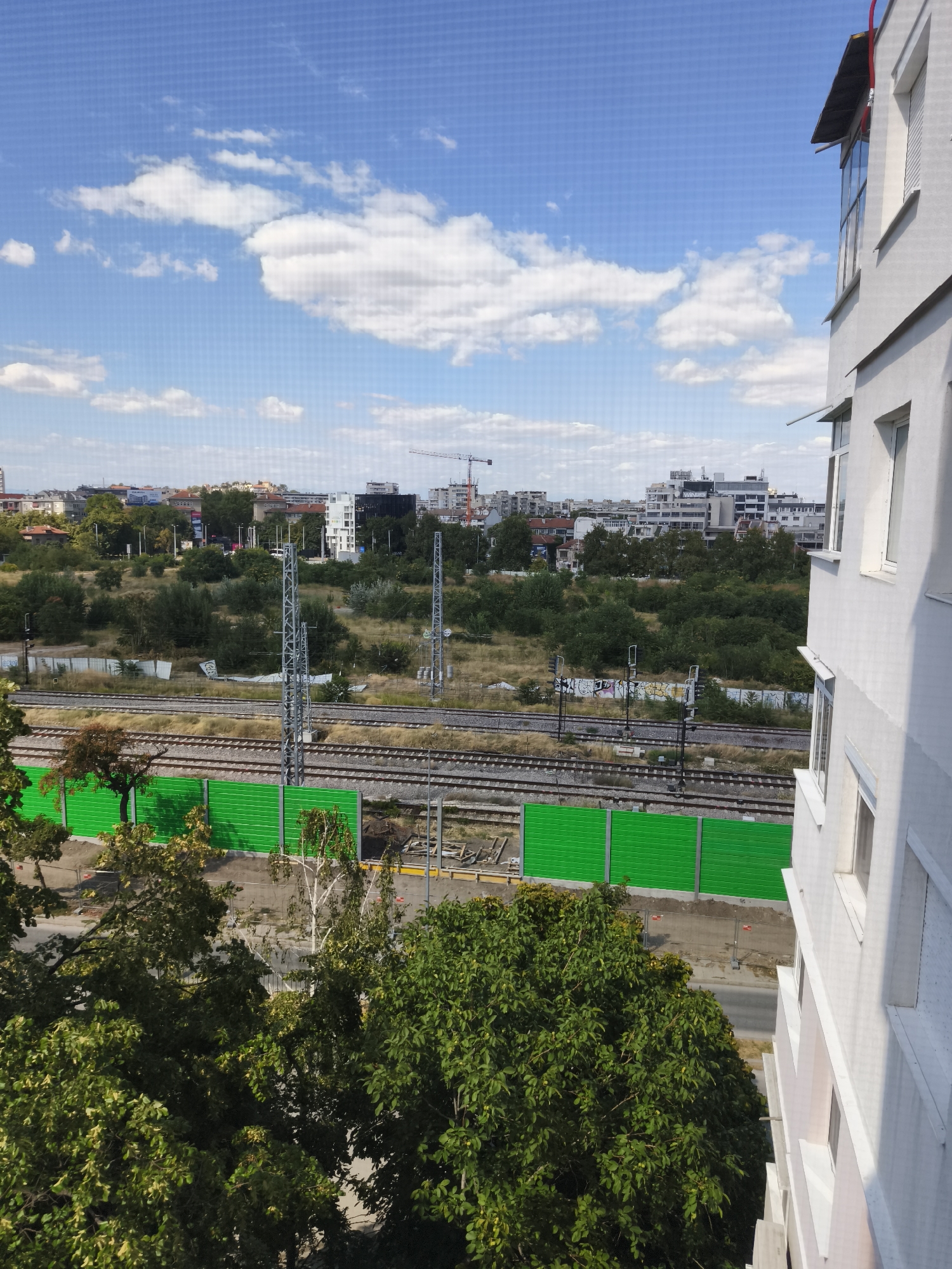 The image depicts an urban scene from an elevated viewpoint, possibly from a building's balcony or window. In the foreground, lush green trees partially obscure the view. To the right, the side of a white, multi-story building is visible, featuring several windows. Beyond the trees and building, a set of train tracks runs horizontally across the frame, separated from the foreground by a bright green sound barrier. In the distance, a cityscape unfolds with various buildings of different heights and architectural styles, interspersed with more greenery. The sky above is a clear blue, dotted with scattered white clouds. Construction cranes are visible in the far distance, suggesting ongoing development in the area. The overall impression is of a residential or mixed-use urban environment.