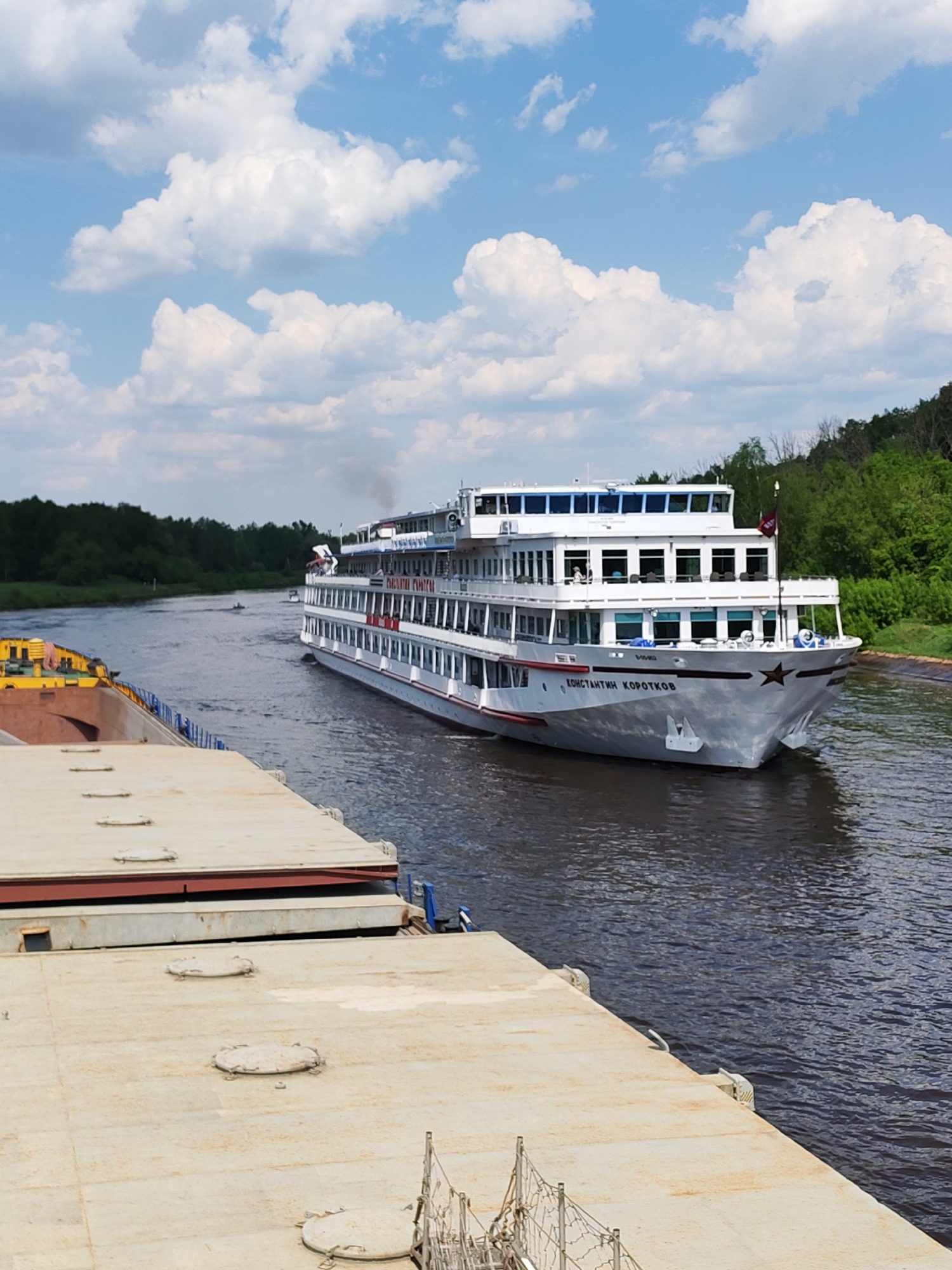 A river scene features a multi-decked, white passenger ship labeled "КОНСТАНТИН КОРОТКОВ" sailing on a waterway flanked by lush green trees. A second watercraft, consisting of yellow and neutral-colored barges, is positioned in the foreground. The sky above displays a mix of blue with scattered white clouds. A small boat can be seen in the distance. The location is Unknown, Unknown.