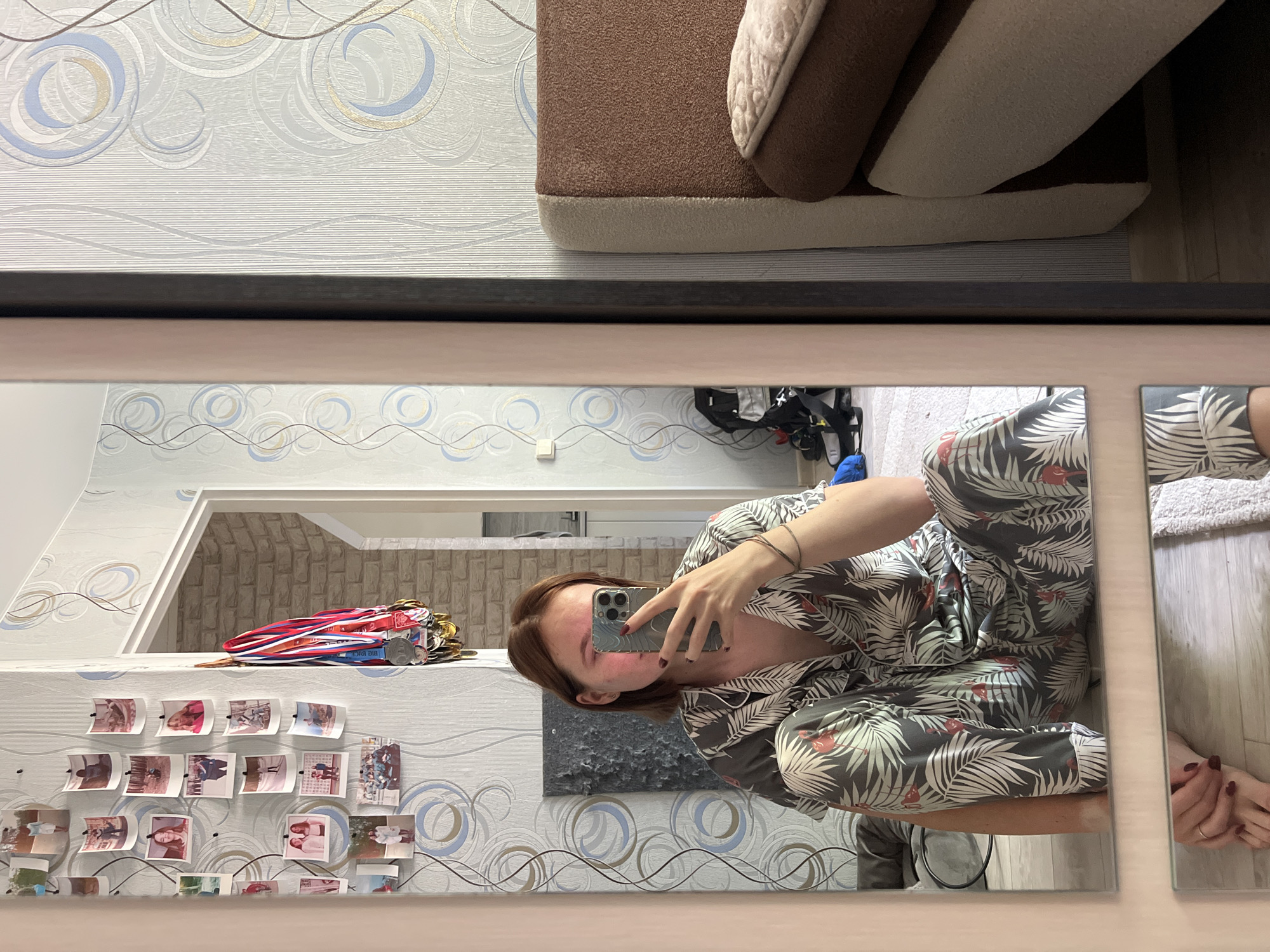 In a bedroom in Chaikovsky, Russia, a young woman with short, light brown hair is sitting on the floor, taking a selfie in a mirror. She is wearing a gray pajama set with a tropical leaf and flamingo print. The room has light blue and white patterned wallpaper, and a collection of framed photos and medals hangs on the wall. To the right of the woman, a portion of a brown and cream-colored headboard of a bed is visible. The floor is wooden, with a rug underneath the woman.