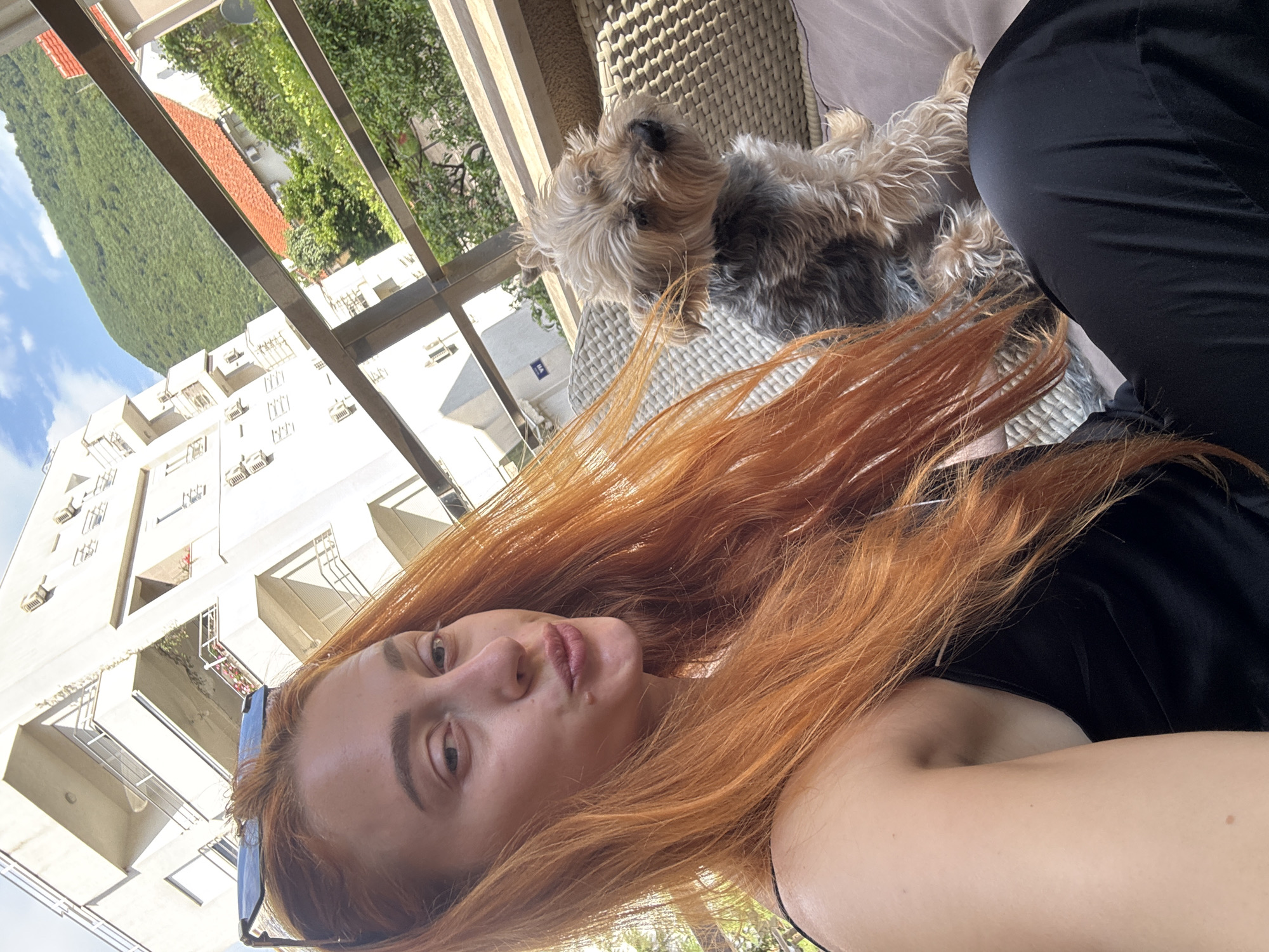 A woman with long, red hair is taking a selfie on what appears to be a balcony or patio. She is wearing a black top. A small, furry dog, possibly a Yorkshire Terrier, is next to her on a wicker-style seat. In the background, there is a multi-story white building with balconies, suggesting an urban residential area. Beyond the building, there's a green, tree-covered hillside and a patch of blue sky. The location is Budva, Montenegro.