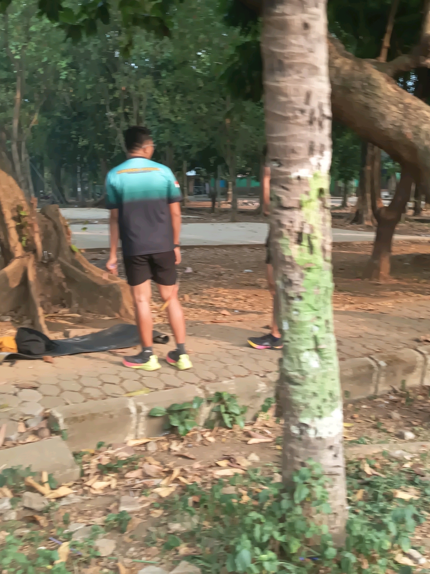 A person is standing on a paved walkway in a park-like setting. The person is wearing a dark blue and teal t-shirt, black shorts, and bright yellow and pink athletic shoes. The person is facing away from the camera. In the foreground, there is a large tree trunk with green and brown coloring. The background shows more trees and some cleared ground. A black bag and a yellow object are lying on the ground near the walkway.