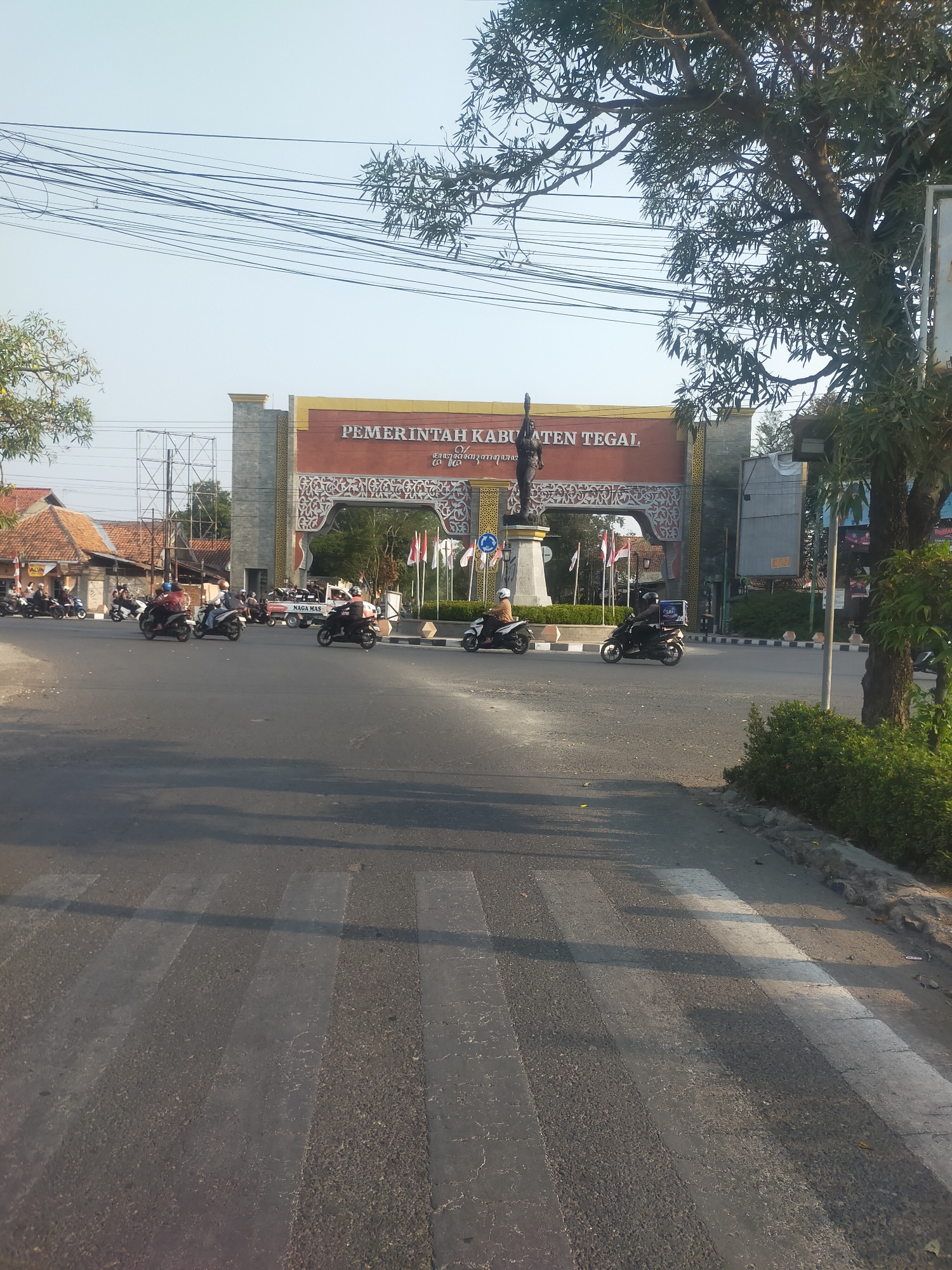 The image depicts a street scene in Kalisapu, Indonesia, in front of the entrance to the "PEMERINTAH KABUPATEN TEGAL" (Government of Tegal Regency). The entrance features a large archway with ornamental carvings and text. A statue of a person raising an arm is prominently displayed on a pedestal within the archway. Several motorcycles are present on the road, with riders in motion. Some motorcycles have packages or bags, suggesting delivery services. The road has a pedestrian crossing marked by white stripes. A large tree with dense foliage is visible on the right side of the image, casting shadows on the road. Overhead, numerous electrical wires crisscross the sky. The weather appears to be sunny with a clear sky. The environment suggests a typical urban or suburban setting in Indonesia with active vehicular traffic.
