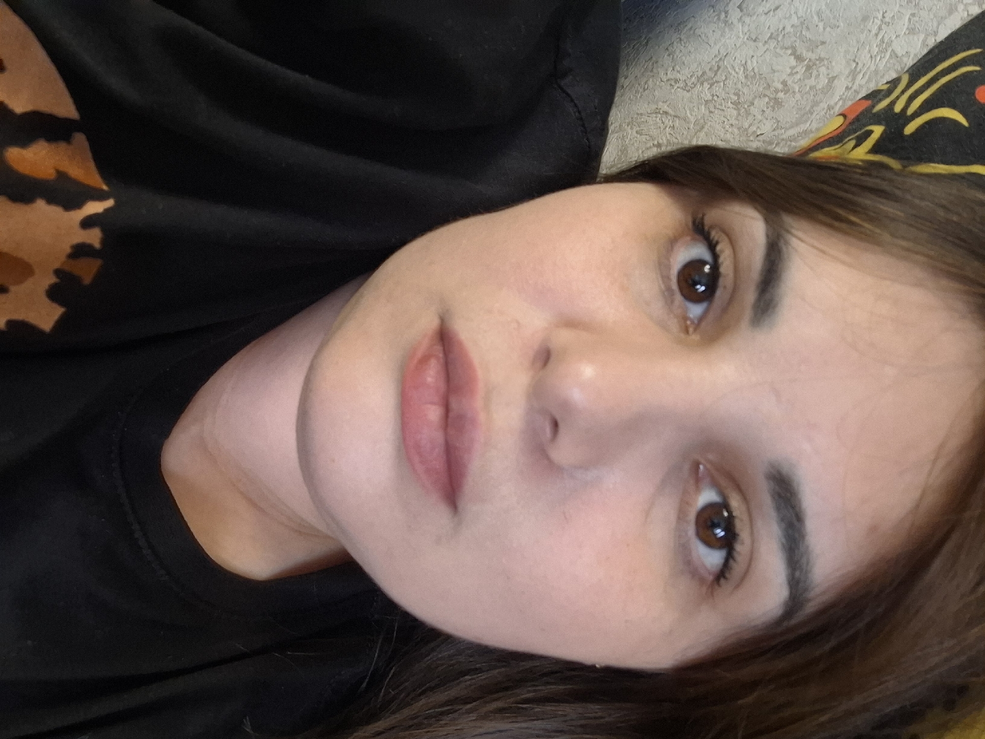 A close-up portrait features a young woman with fair skin, dark brown hair, and dark brown eyes, looking directly at the camera. She appears to be lying down, with the image taken from an elevated perspective, tilting her head slightly. Her expression is neutral and calm.
She is wearing a black t-shirt or top, part of which displays an abstract, multi-toned pattern in shades of brown and orange, reminiscent of an animal print (possibly leopard or cheetah). Her lips are softly colored with a pinkish-nude hue. Her eyebrows are well-defined, and her eyelashes appear enhanced.
Behind her head, to the right side of the frame, there is a visible section of patterned fabric, featuring yellow and red shapes on a dark background, suggesting a pillow or blanket. The background behind her body appears to be a textured, light-colored surface, possibly a wall or upholstery.
There is no discernible activity taking place other than posing for the photograph. No text is visible in the image. The image provides an intimate, indoor setting. While the location context is Chaikovsky, Russia, no specific visual cues within the image itself confirm this.