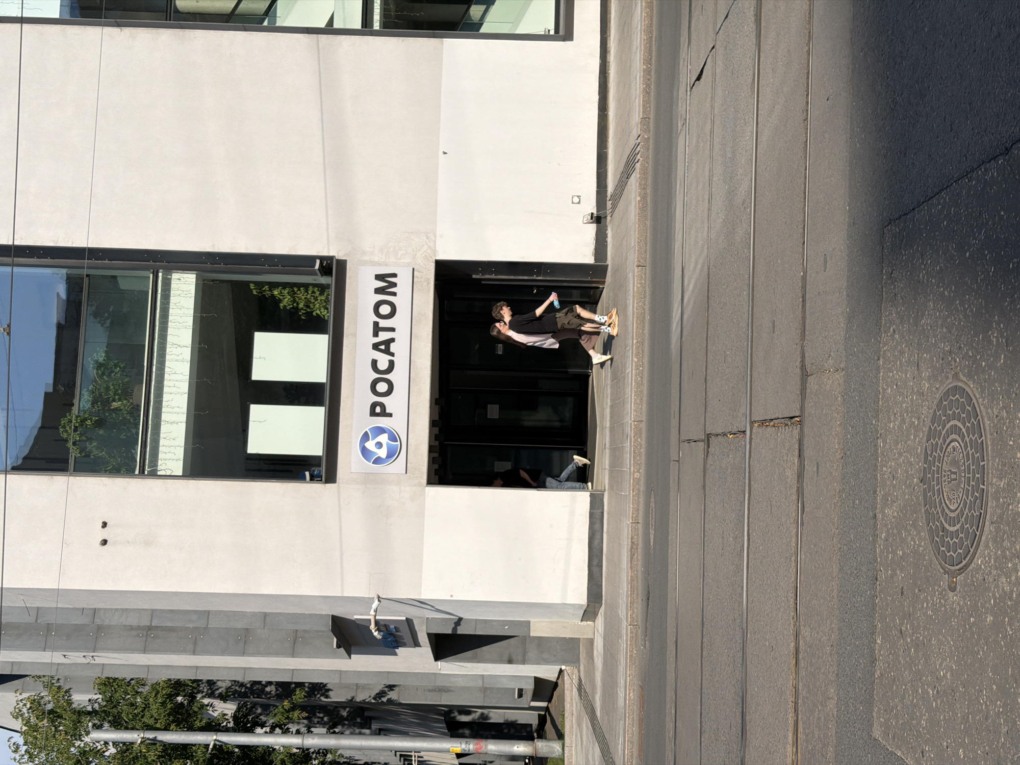 The image shows a street-level view of the entrance to a building with "POCATOM" signage. The building appears to be modern, with a grey concrete facade and large glass windows. In front of the entrance, a set of dark glass doors is visible. Two individuals are walking out of the building, one slightly ahead of the other. The person in front, wearing a black t-shirt and olive-colored shorts, is looking down and holding a blue bottle. The second person, a woman wearing a light pink t-shirt and dark pants, is walking beside them. The street in the foreground shows tram tracks, indicating it is a public thoroughfare. The overall environment suggests an urban setting, likely a business district. The location is specified as the city of Moscow, Russia.