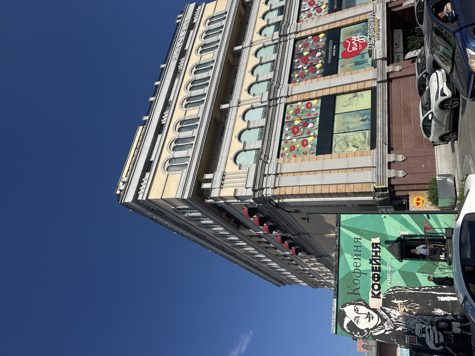 In Yekaterinburg, Russia, a low-angle shot captures the facade of a building under a clear blue sky. The building, with its pale yellow and grey architecture, features multiple levels of arched windows and ornate detailing. Red awnings extend from some of the upper windows. Below, the ground floor has large display windows showcasing floral patterns and advertisements.
To the left of the main building, a smaller, green structure is prominently displayed. This structure features a large black and white mural of a man holding a coffee cup, with text above in Cyrillic, including "Кофейня" which translates to "Coffee House." Two people are visible near the entrance of this smaller building, one standing on the steps and another partially obscured by the mural.
In front of the buildings, several cars are parked on the street, including a white SUV and a dark blue car. The overall scene suggests a commercial district with a mix of architectural styles and urban activity.