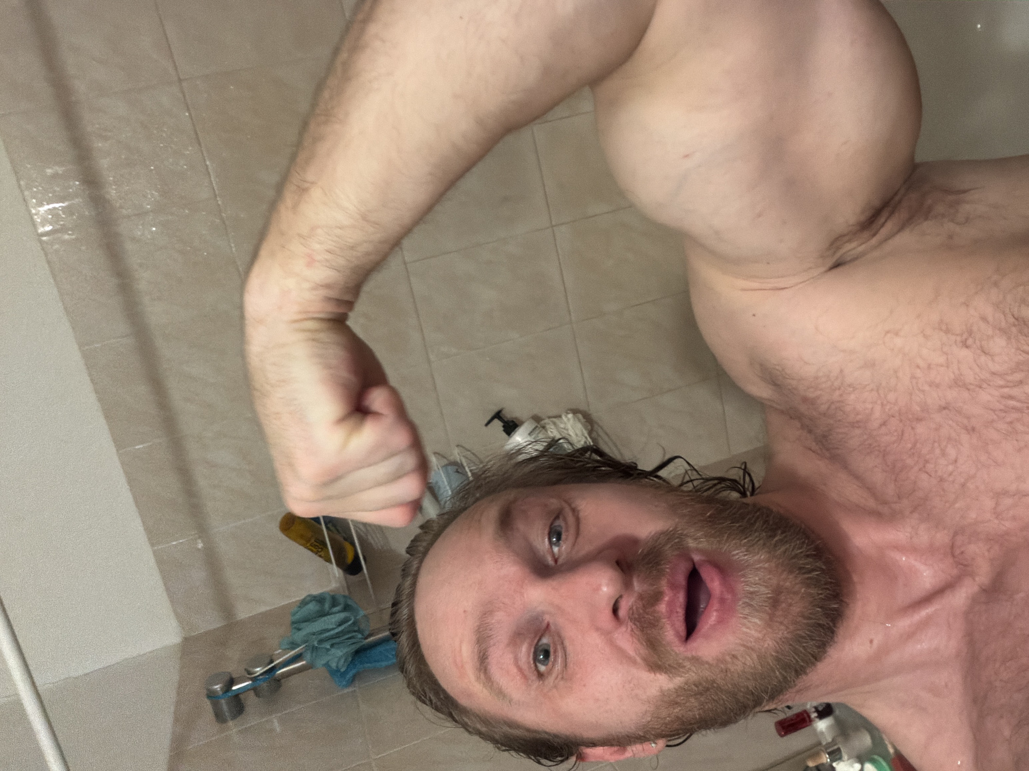 The scene depicts a male individual taking a selfie within a residential bathroom, identified as being in Saint Petersburg, Russia. The man, with wet reddish-brown hair and a beard, is shirtless and appears to be wet from showering. He looks directly at the camera with wide blue eyes and an open mouth, conveying a sense of surprise or excitement. His right arm is flexed, prominently displaying a muscular bicep towards the upper right of the frame. The background consists of light-colored, rectangular tiled walls, typical of a shower enclosure. A chrome shower caddy or organizer is mounted on the left wall, holding a blue loofah, a yellow cylindrical bottle, and a white pump-top bottle. A white shower rail is visible across the top of the frame. Water droplets are visible on the man's skin and potentially on the tiled surfaces.