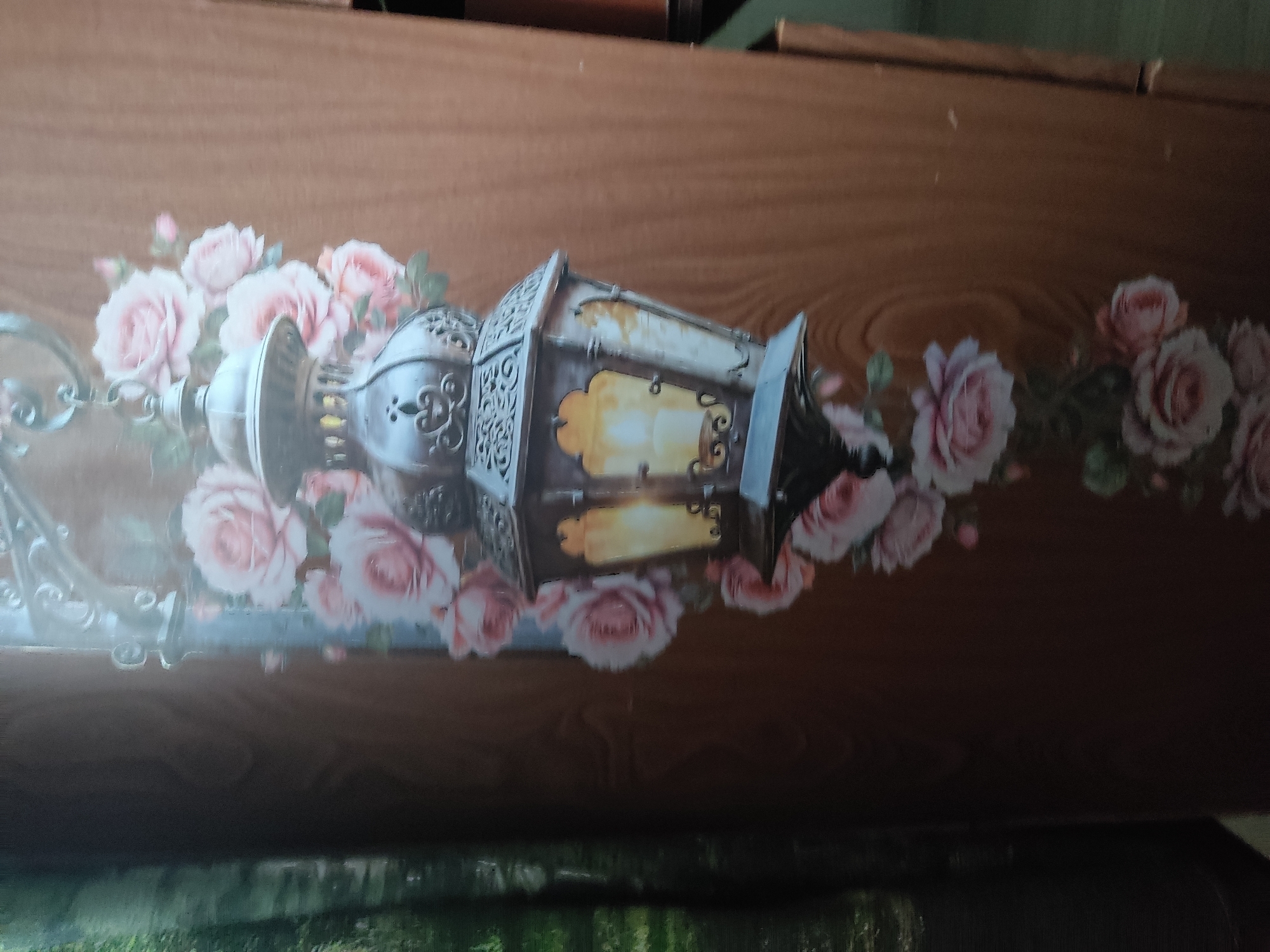 A photograph shows a close-up view of a wooden surface with a decorative decal. The decal features a vintage-style lantern surrounded by pink roses. The lantern is ornate, with intricate metalwork and glass panes that appear to glow with a warm light. The roses are depicted in various stages of bloom, with soft pink petals and green leaves. The wooden surface has a natural grain pattern and a warm, brown hue. The lighting in the image is soft and diffused, highlighting the detail of the decal. There are no people or animals visible in the photograph. The context of this image in Noginsk, Russia, is unknown.