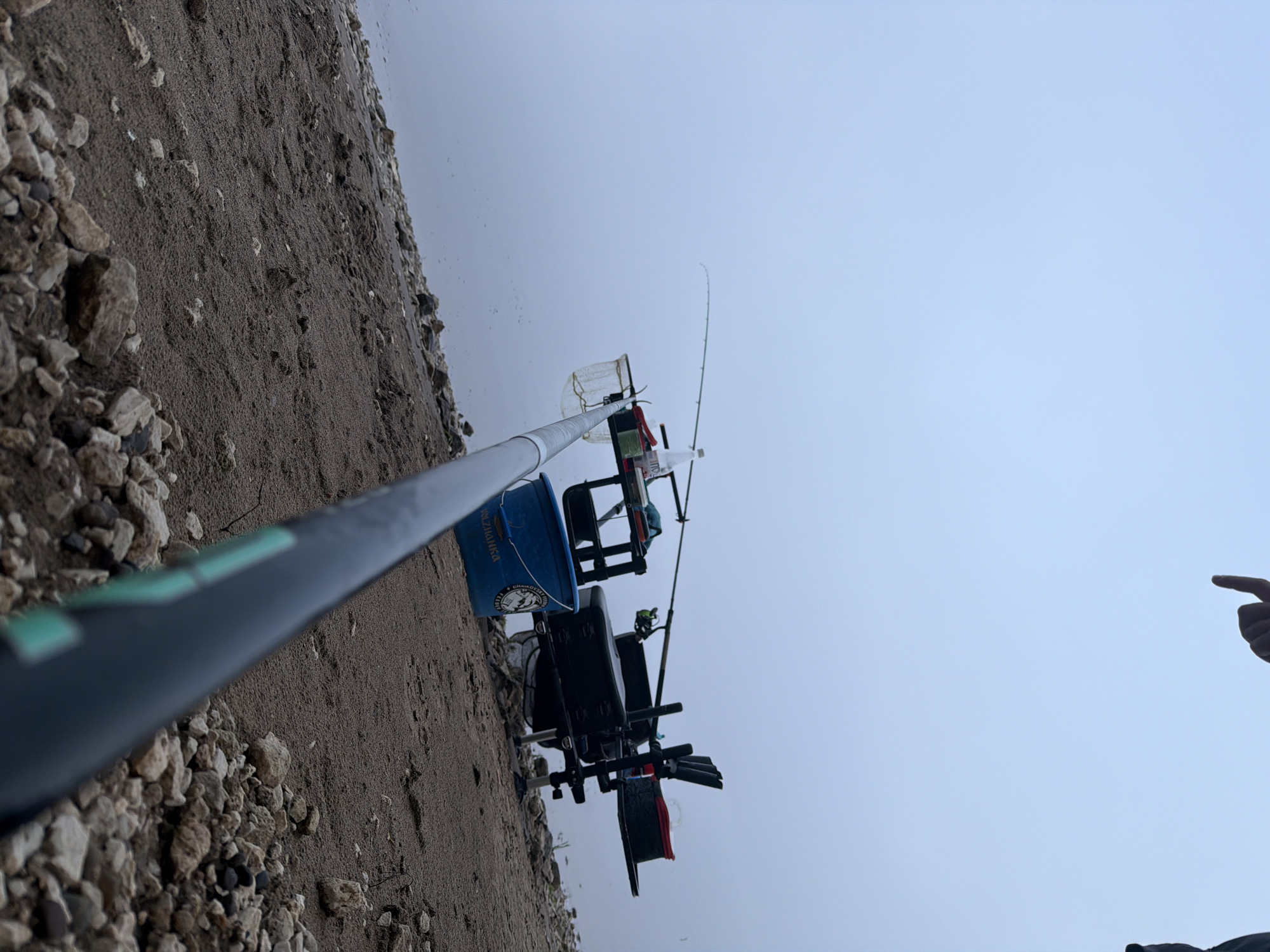 The image presents a low-angle view of a fishing setup on the rocky and sandy shore of a body of water, likely a riverbank in Chaikovsky, Russia. The central activity is fishing, with equipment meticulously arranged by the water's edge under an overcast sky.
In the midground, a black fishing chair or platform is positioned, equipped with multiple rod holders and other attachments. To the left of the chair, a blue bucket is visible, emblazoned with the Cyrillic text "ВОЛЖАНКА" and its Latin transliteration "VOLZHANKA," alongside a logo featuring a fish. A white bottle also rests near the setup. Two fishing rods are present: one, partially blurred, extends from the bottom left corner across the foreground, while a second, thinner rod is set up further back, pointing over the calm, reflective water. A white fishing net on a pole stands ready behind the main fishing station.
The ground comprises a mix of light-colored gravel and darker, finer sand or dirt. The water surface is smooth and reflects the uniform, light gray-blue of the sky, indicating a cloudy or hazy day. On the far right of the frame, a human hand with an index finger pointing upwards is partially visible against the sky. The scene conveys a sense of quiet anticipation associated with recreational fishing.