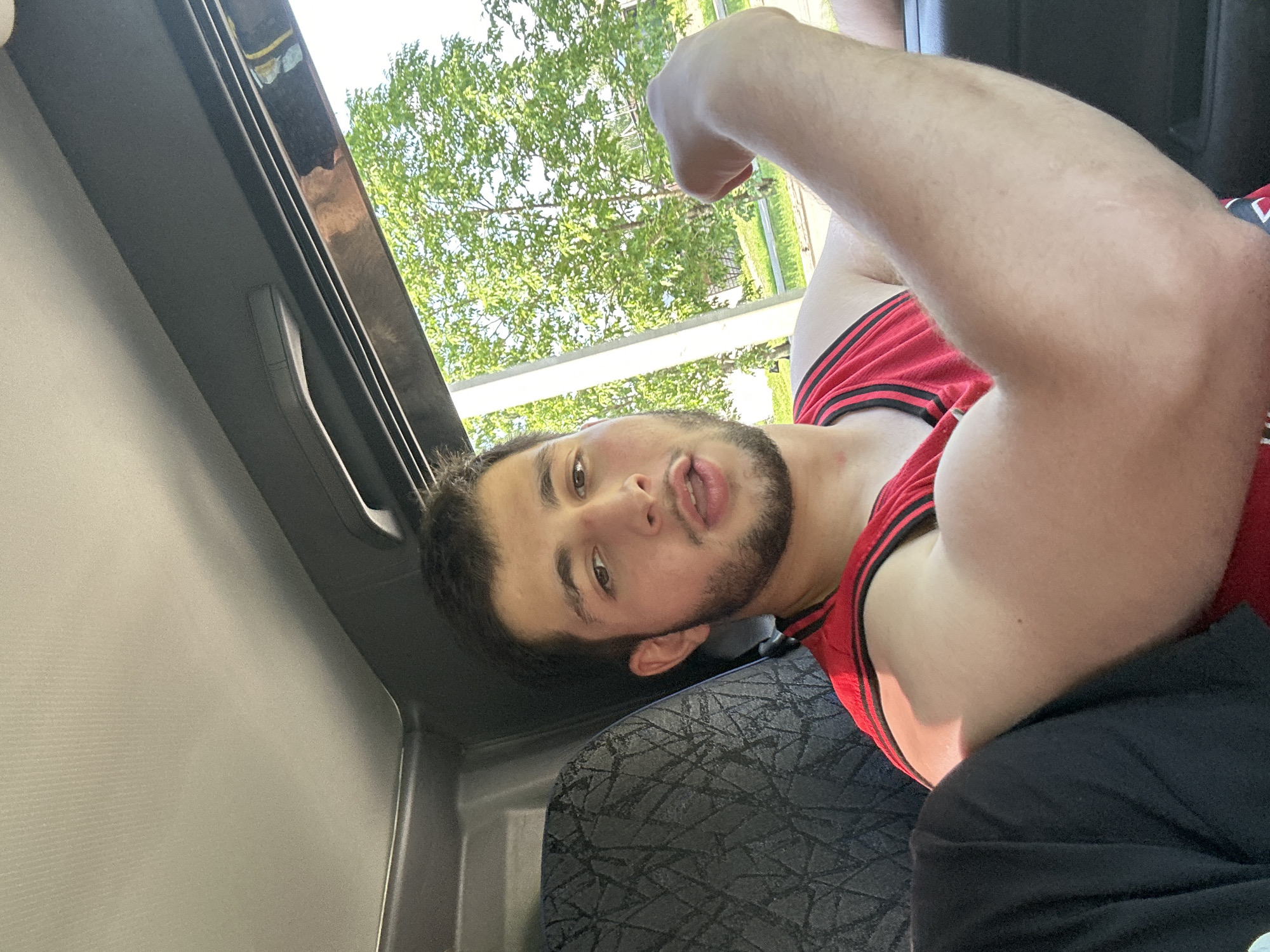 A young man with dark hair and a beard is sitting in the passenger seat of a vehicle, looking at the camera with his mouth slightly open. He is wearing a red tank top with black trim. His right arm is prominently displayed, flexed, and showing his bicep. The interior of the vehicle is visible, including a black textured seat and the car's ceiling. Outside the window, green trees and a clear sky are visible, suggesting an outdoor setting, possibly on a bright day. The image appears to be a selfie taken within the car. No specific location in Birobidzhan, Russia is identifiable from the visual information.