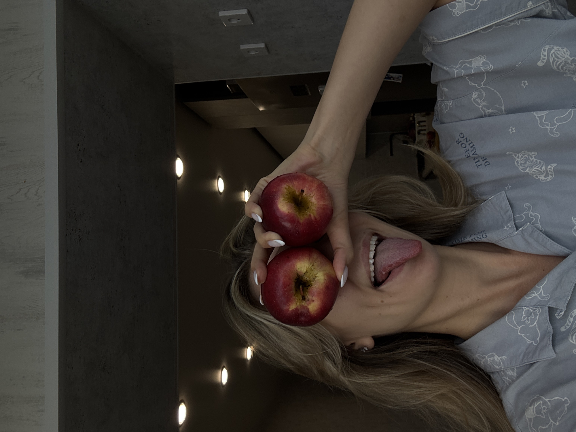 The image, rotated clockwise, depicts a person playfully posing indoors. The subject, likely a woman with light brown hair, is seen from the chest up, with her head tilted back and her tongue extended from her mouth in a wide, joyful expression. She holds two bright red apples, one in each hand, directly over her eyes, obscuring them. Her hands show well-manicured fingers with white nail polish.
She is wearing light blue pajamas patterned with white outlines of cartoon tigers and stars. The text "TIME FOR DREAMING" is clearly visible on the front of her pajama shirt. A small, possibly silver, earring is visible in her left ear.
The indoor background features a dark, textured wall on the left side of the frame, punctuated by several glowing, circular wall-mounted lights. To the far left, a lighter, possibly concrete or wood-paneled wall is partially visible. The upper portion of the room, towards the top of the rotated image, shows a dark grey ceiling or upper wall with two rectangular fixtures. The lighting is dim except for the accent lights. The overall mood conveyed is one of playfulness and humor.