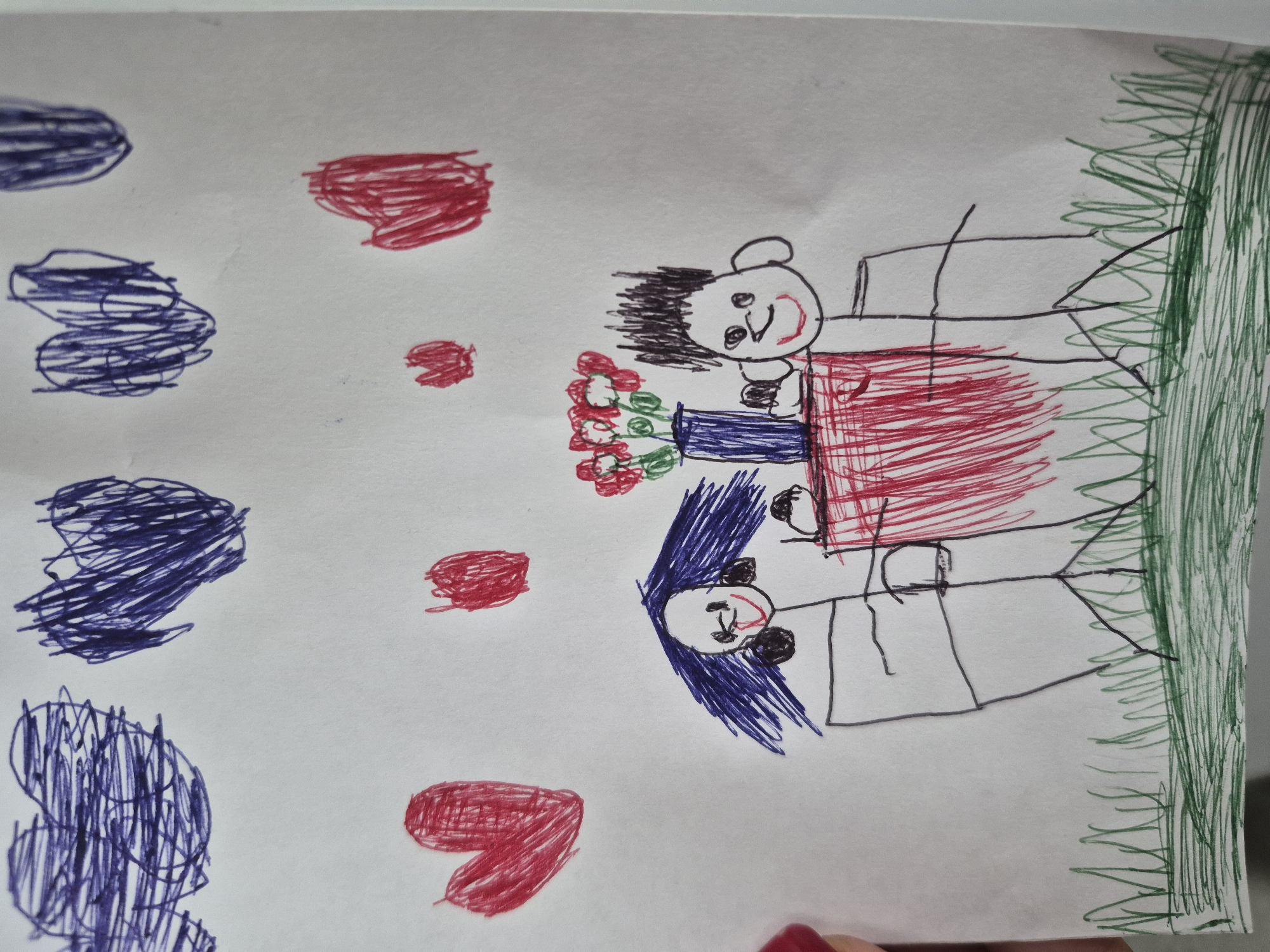 The image is a child's drawing. It depicts two figures, presumably people, sitting at a table. A vase with red flowers is on the table between them. The figures appear happy. There are red hearts and blue shapes drawn around the figures. Green scribbles are on the right, possibly indicating grass or foliage. There is no indication of the city of Tobolsk, Russia, in the image.