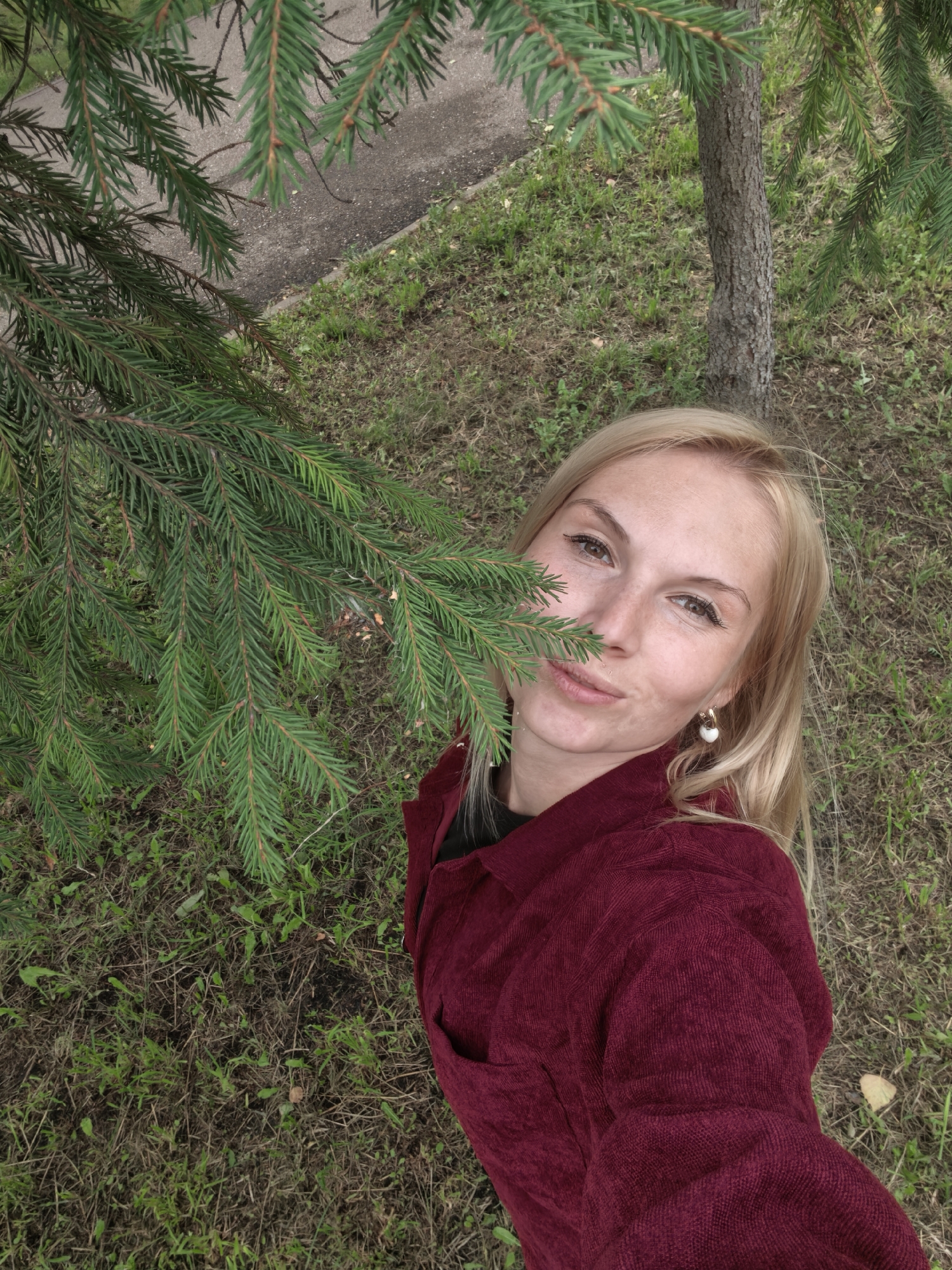 A woman with blonde hair and fair skin is taking a selfie outdoors, partially obscured by a spruce tree branch. She is wearing a dark red corduroy shirt and a white earring in her left ear. The background consists of grass, a tree trunk, and a paved surface. The scene appears to be a park or a similar outdoor area.