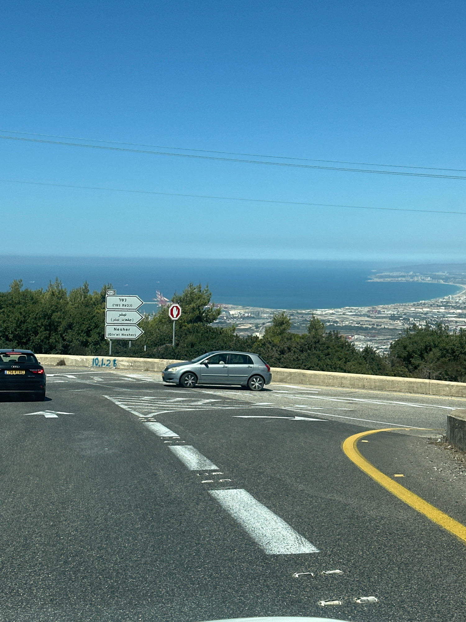 The image depicts a view from a road overlooking the city of Nesher, Israel, and the Mediterranean Sea. In the foreground, a paved road curves to the right with white lane markings and a yellow dividing line. Two cars are visible on the road: a black car on the left and a silver hatchback on the right. A signpost with directions in Hebrew and English points towards Nesher, specifically mentioning "Nesher (Giv'at Nesher)". Trees and vegetation line the side of the road. In the background, the cityscape of Nesher and the port area spread out towards the blue sea under a clear sky. The overall scene captures a moment on a highway with a scenic overlook of an Israeli coastal city.